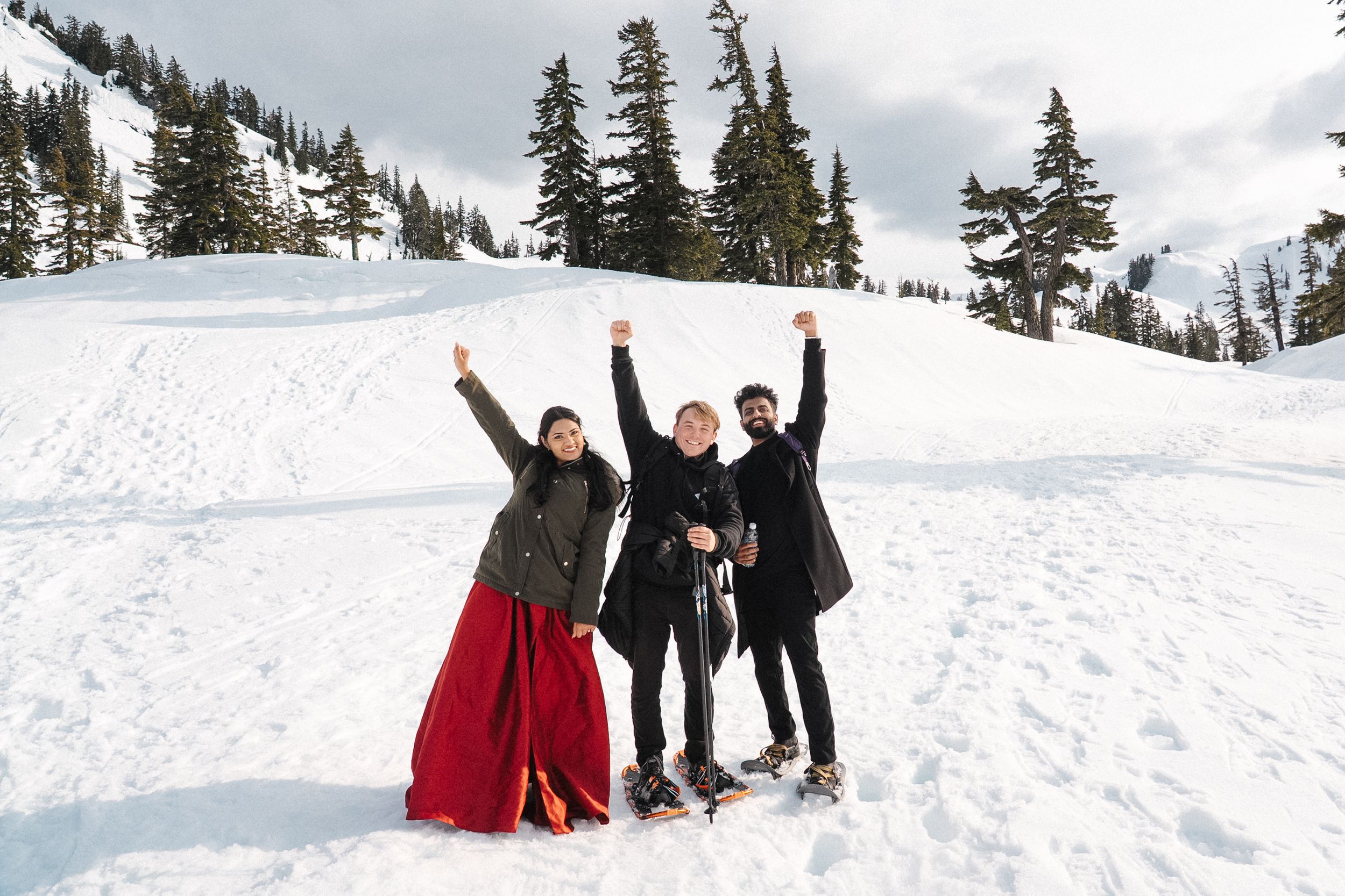 Three friends celebrate joyfully in winter clothes with arms raised on a snowy mountain slope with evergreen trees.