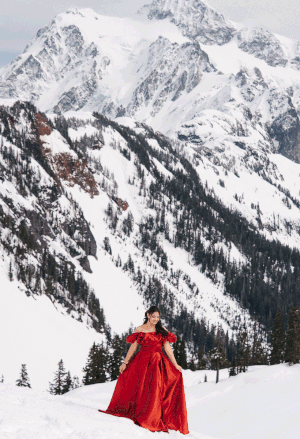 A red ballgown flows gracefully against a snowy mountain backdrop in this stunning winter landscape photograph.