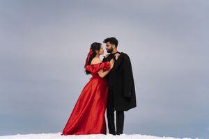 Romantic couple in red dress and black suit embraces against snowy mountain backdrop during winter photoshoot.