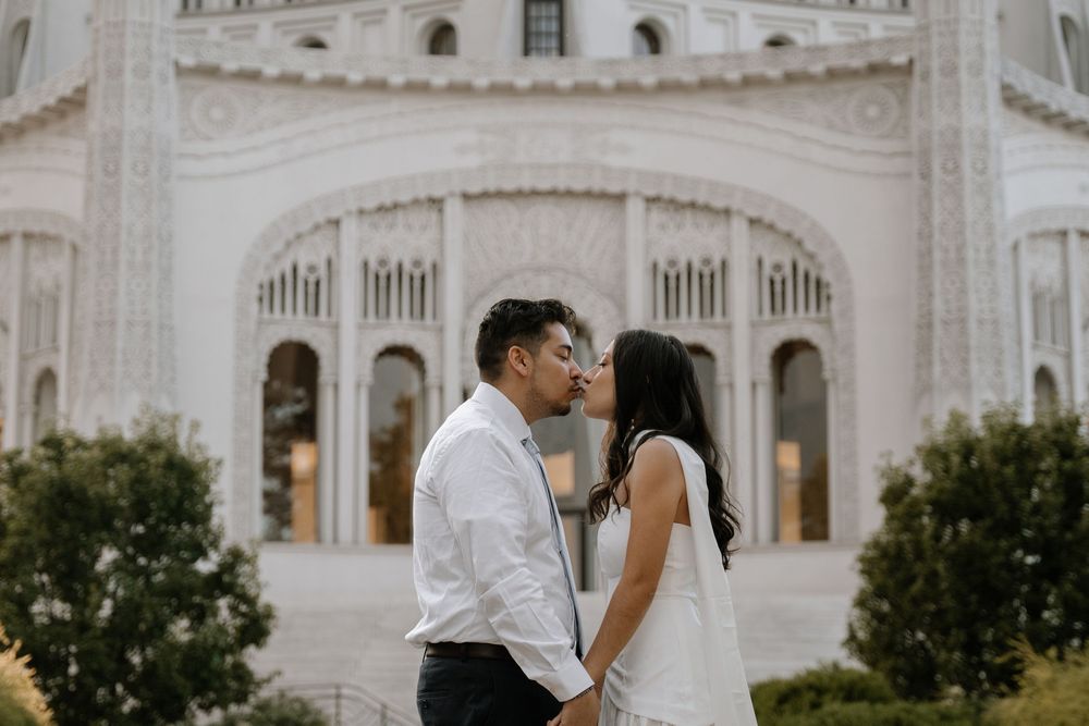 Elegant couple embraces outside ornate white marble temple with arched windows and columns.