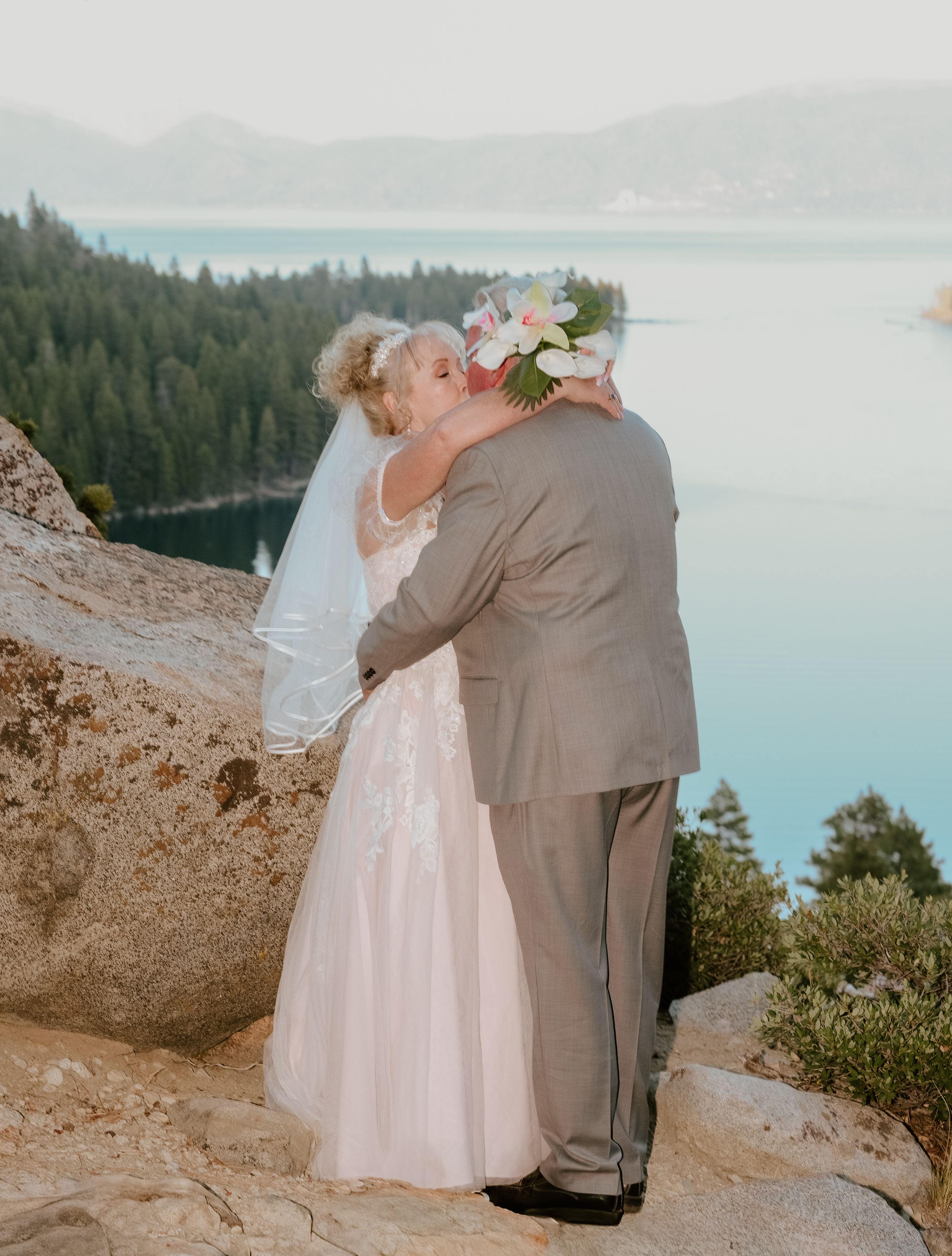 A romantic wedding couple shares tender moments on a cliff overlook with a scenic lake and forest vista in the background.
