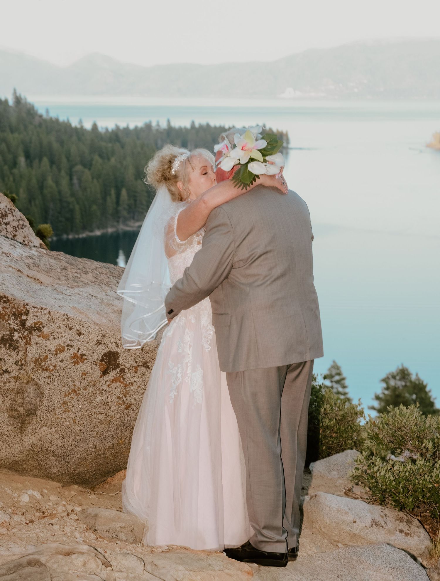 A romantic wedding couple shares tender moments on a cliff overlook with a scenic lake and forest vista in the background.