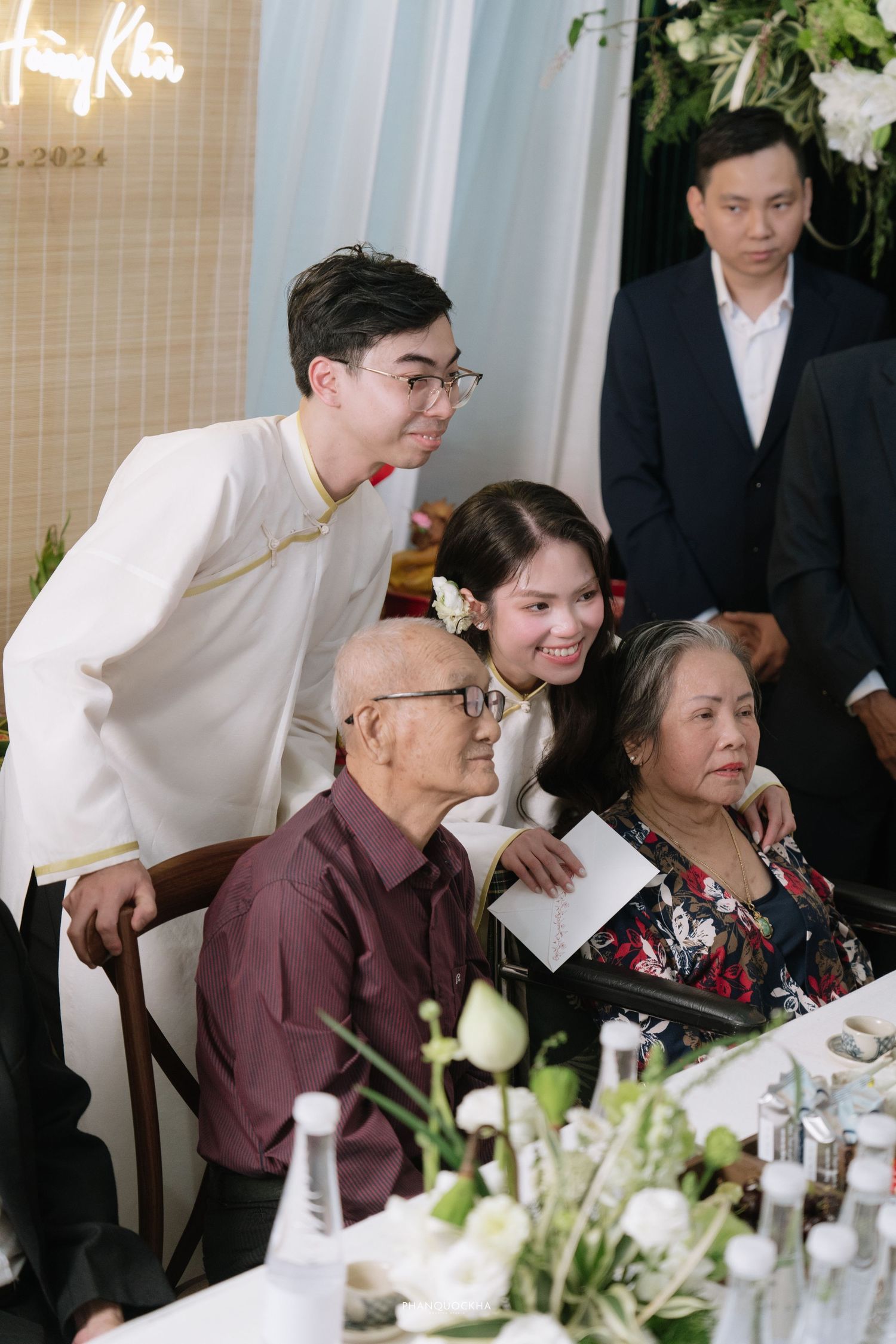 Vietnamese wedding couple in white ao dai pose with elderly grandparents at reception table with white floral arrangements.
