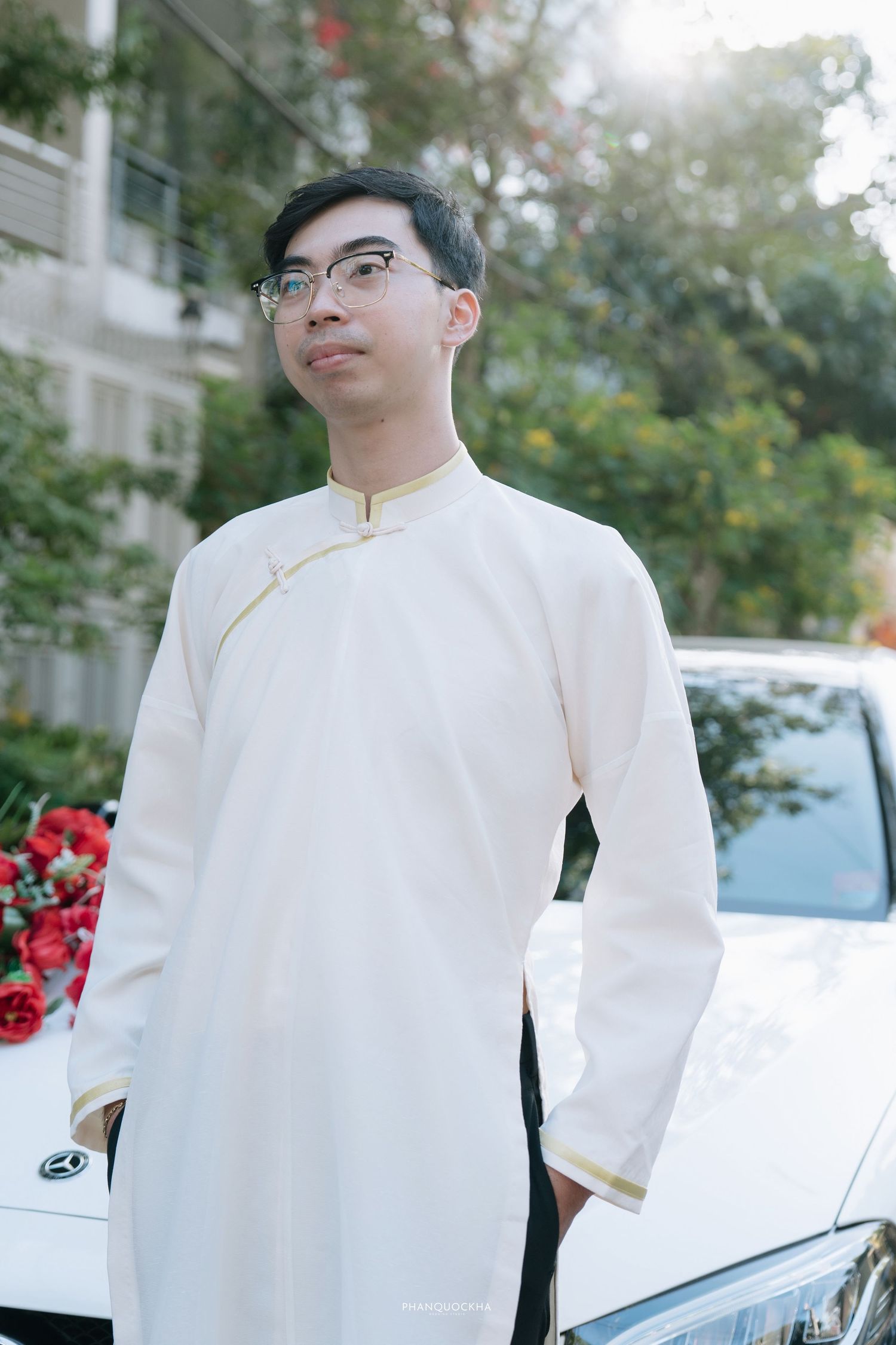 Young Vietnamese groom in white ao dai with glasses standing beside flower-decorated wedding car.