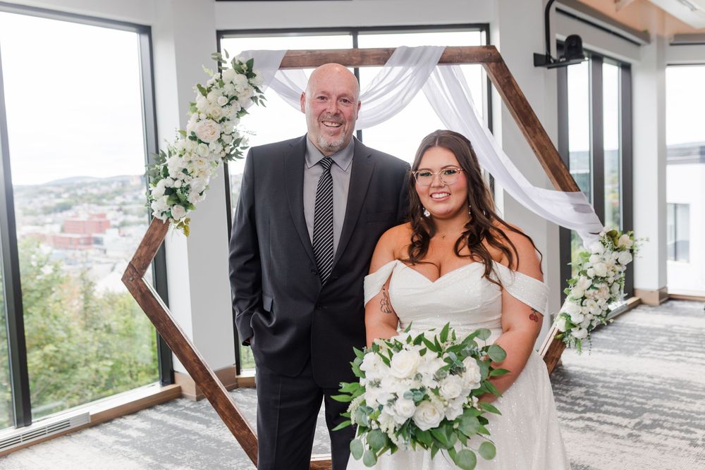 Wedding party poses in a bright, modern indoor venue with white floral arrangements and large windows.