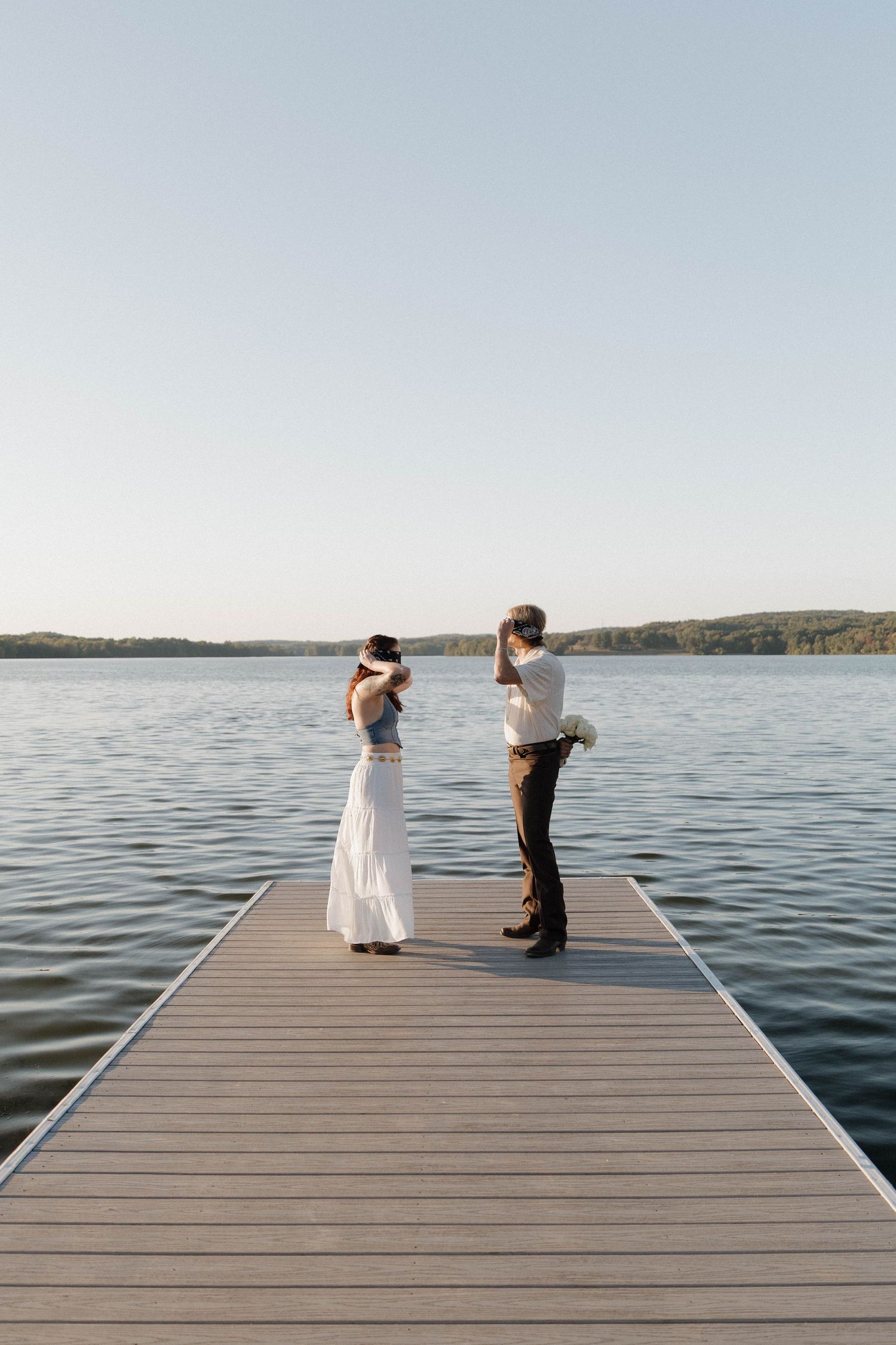 Two people wearing masks stand together on a wooden dock overlooking a serene lake at sunset.