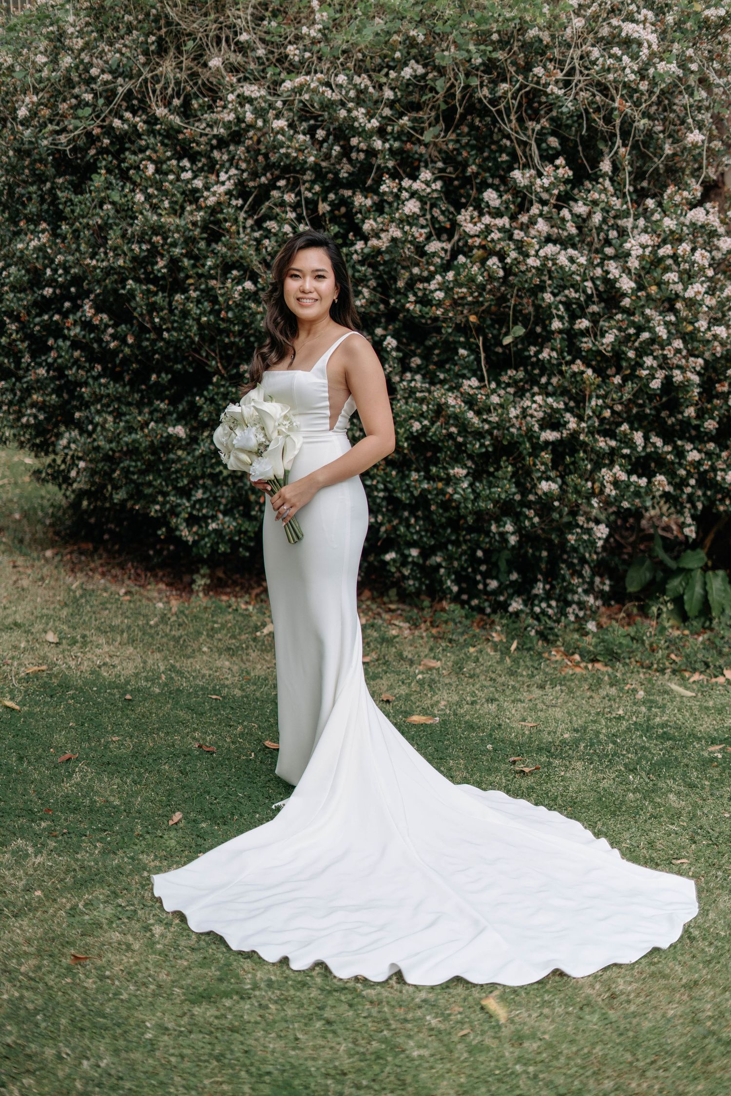 A stunning white wedding dress with a dramatic long train flows gracefully across green grass and garden scenery.