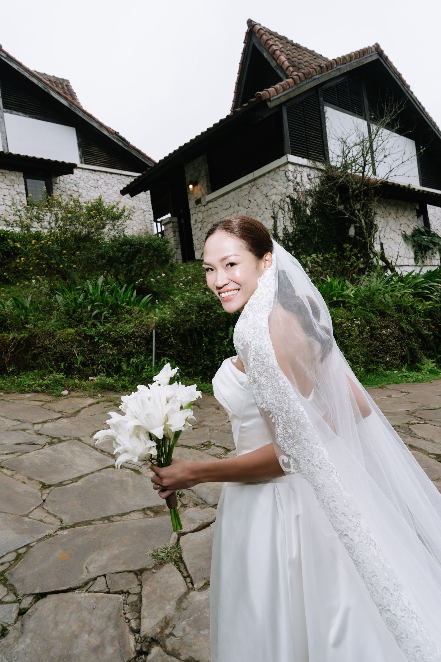 Bride in white wedding dress and veil smiling while holding white floral bouquet outside.