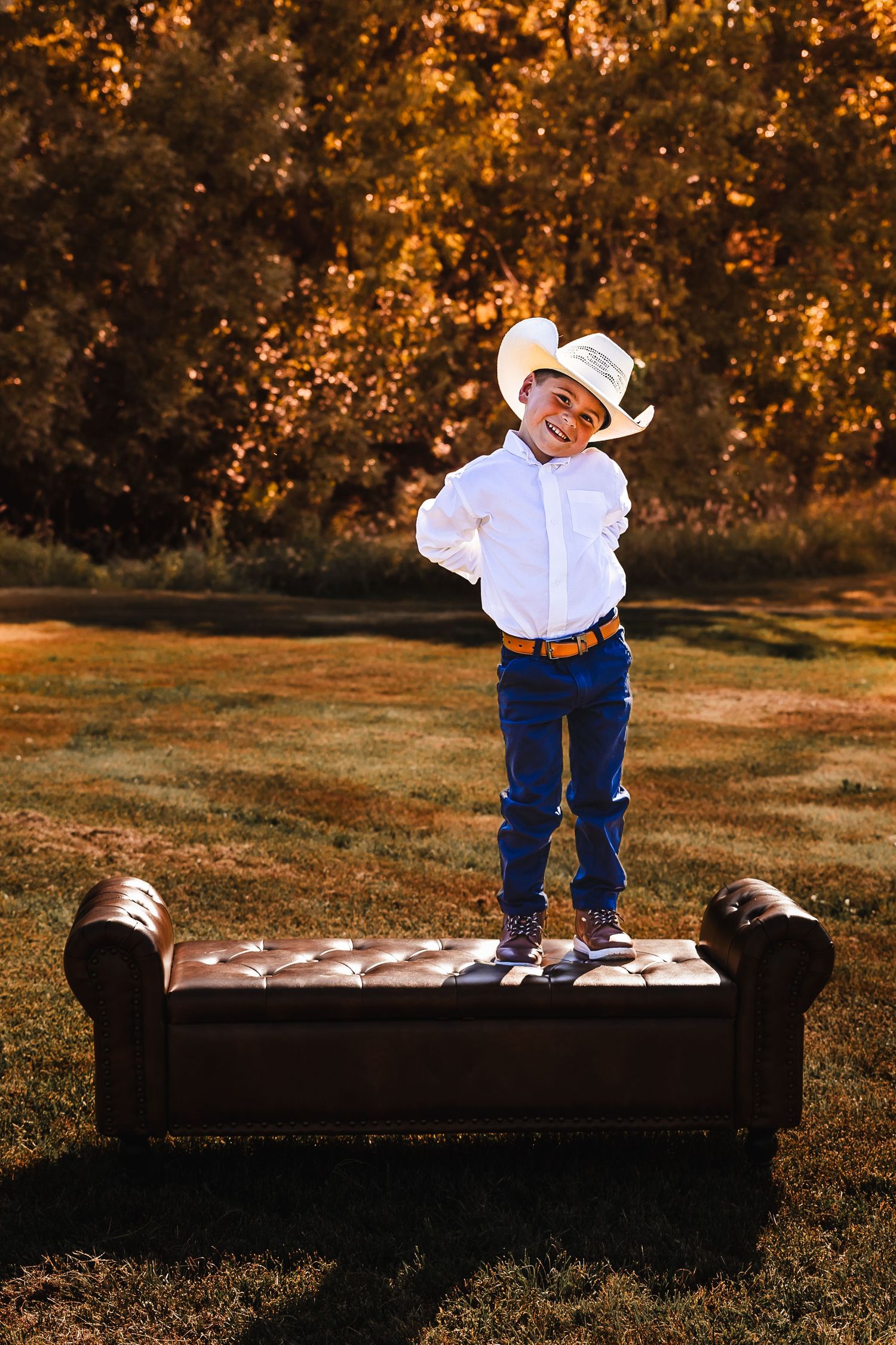 A small child in western wear stands on a leather bench during golden hour with autumn trees in the background.