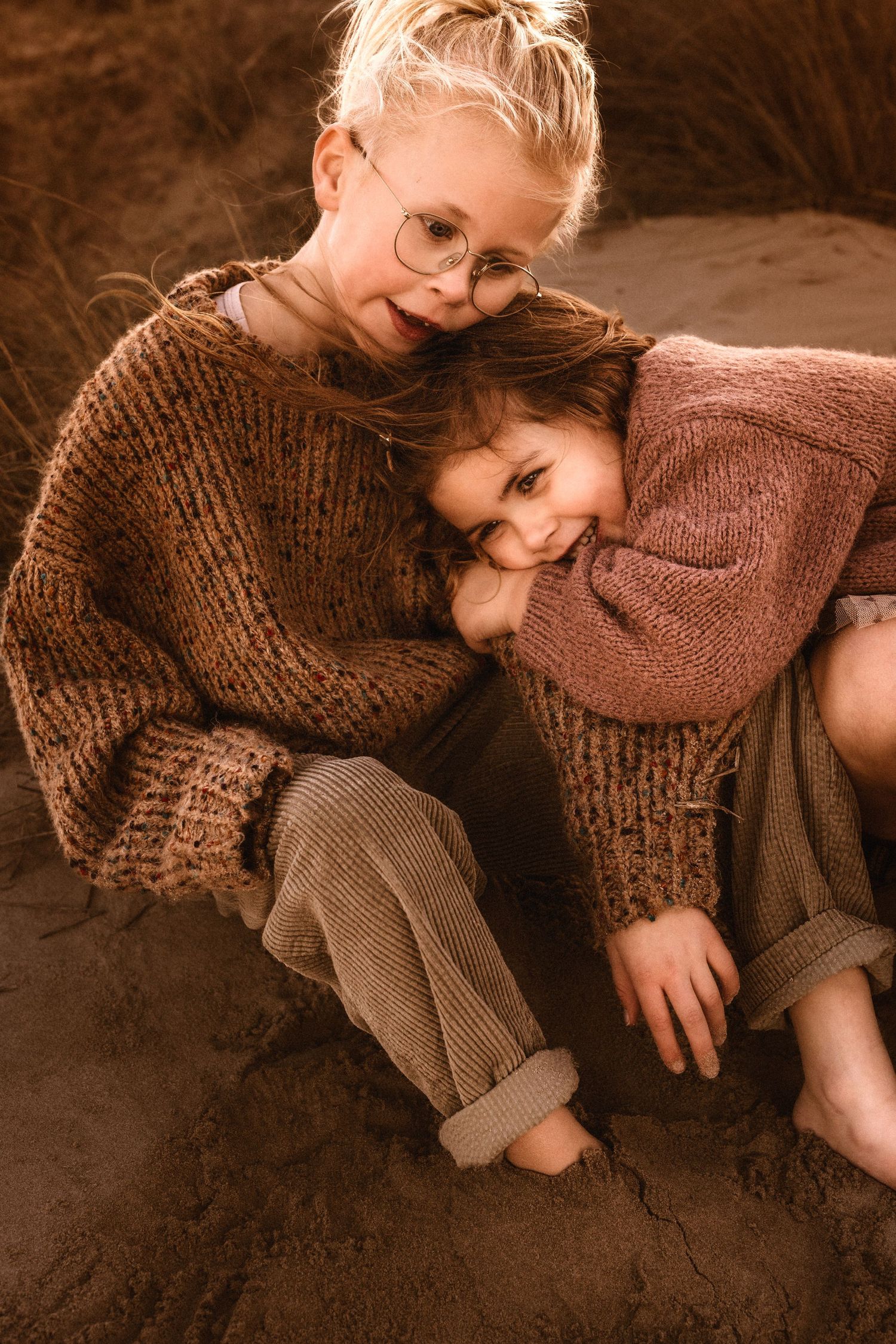Siblings in cozy brown and rust-colored knit sweaters in Denia cuddle together on a dark surface in warm lighting. Denia photographer