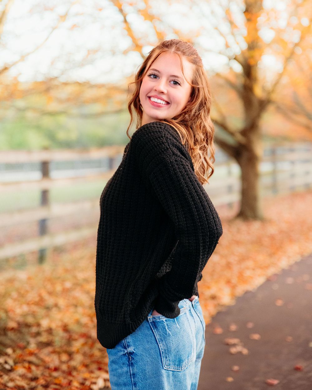 Person in denim jeans and black sweater walks along autumn path with fallen orange leaves and wooden fence.