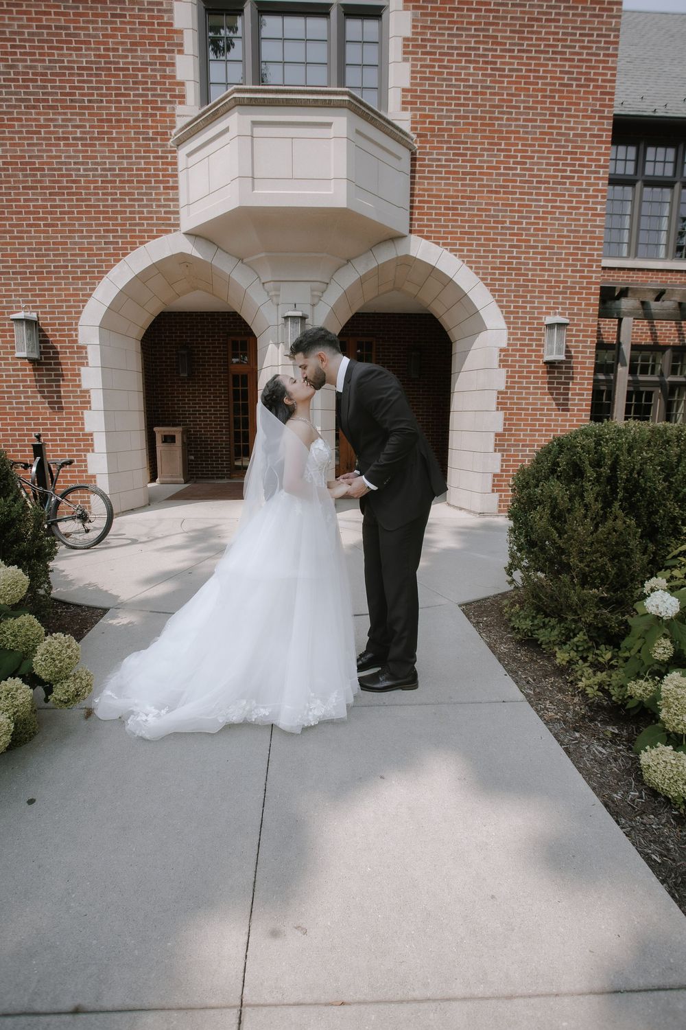Five frame sequence of a couple's first kiss as newlyweds outside a brick building with arched windows.