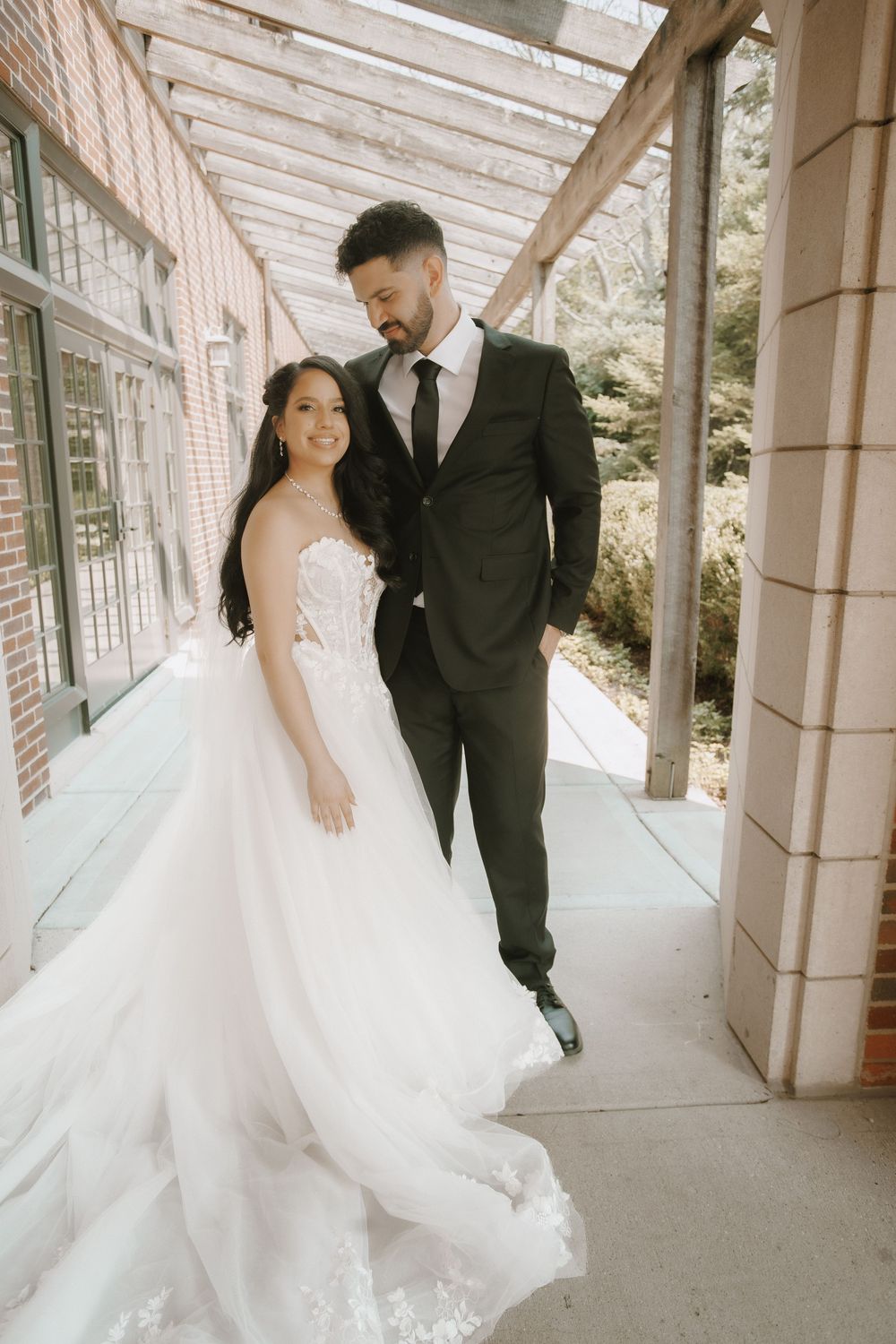 A couple in wedding attire embraces while standing in a columned walkway with brick walls.
