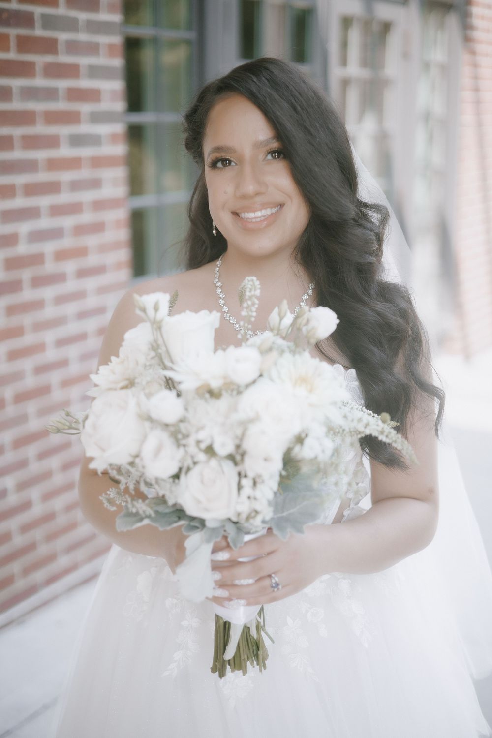 Bride in a white gown holds a luxurious bouquet of white flowers against a brick backdrop.