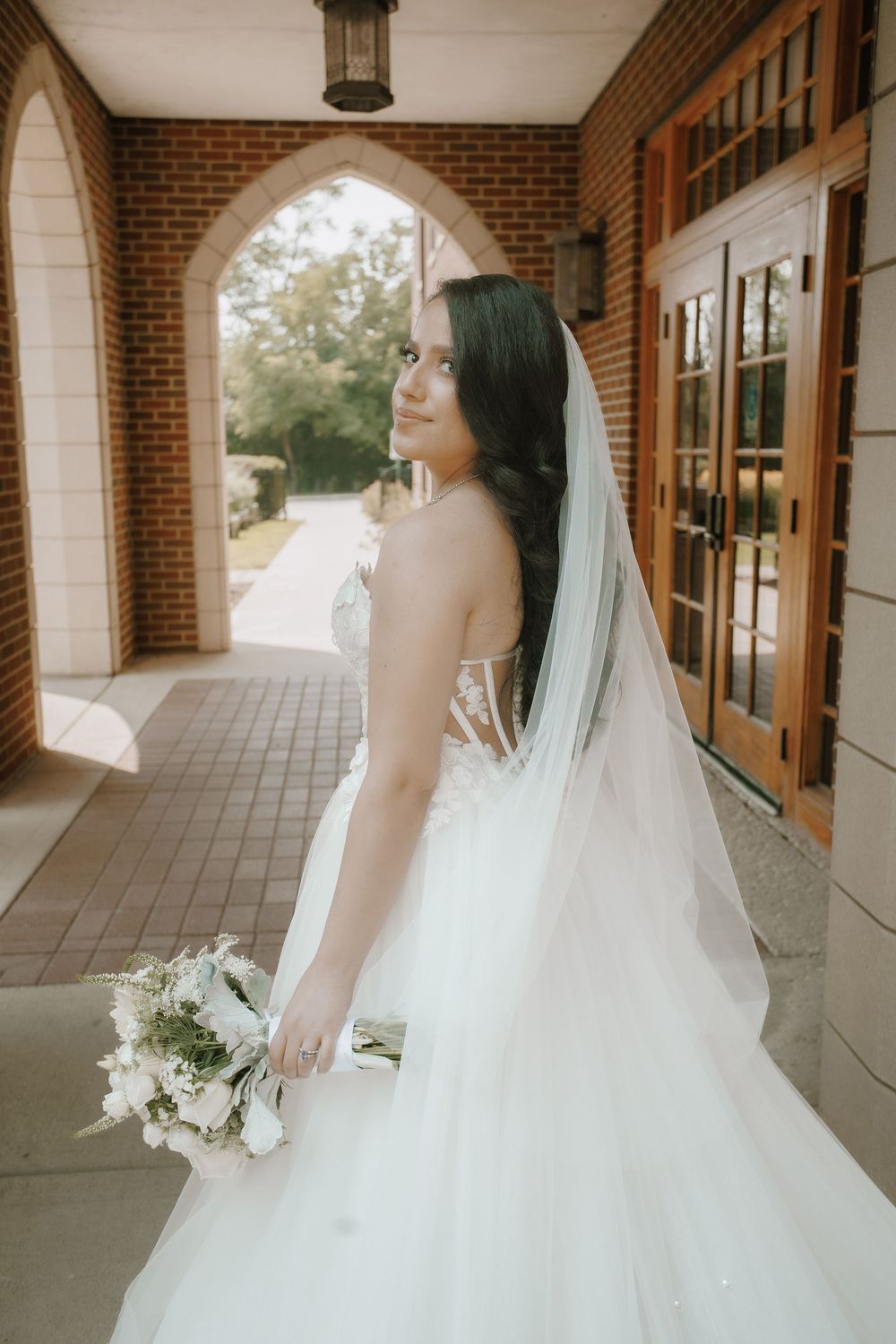 Elegant bride in flowing white ballgown poses along a covered brick walkway with natural lighting and white bouquet.