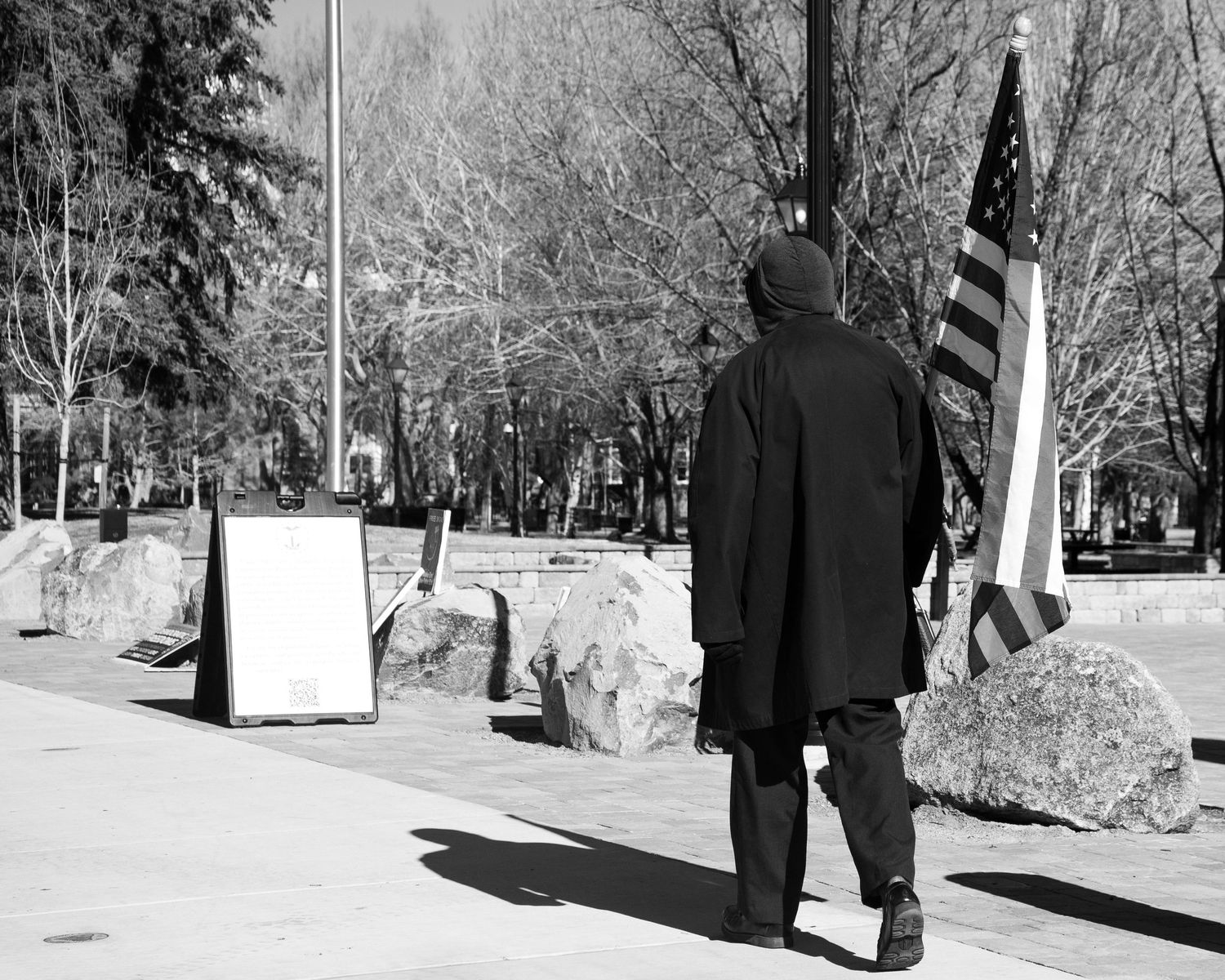 Black and white photo of a hooded figure walking past an American flag and rocks on a city sidewalk.