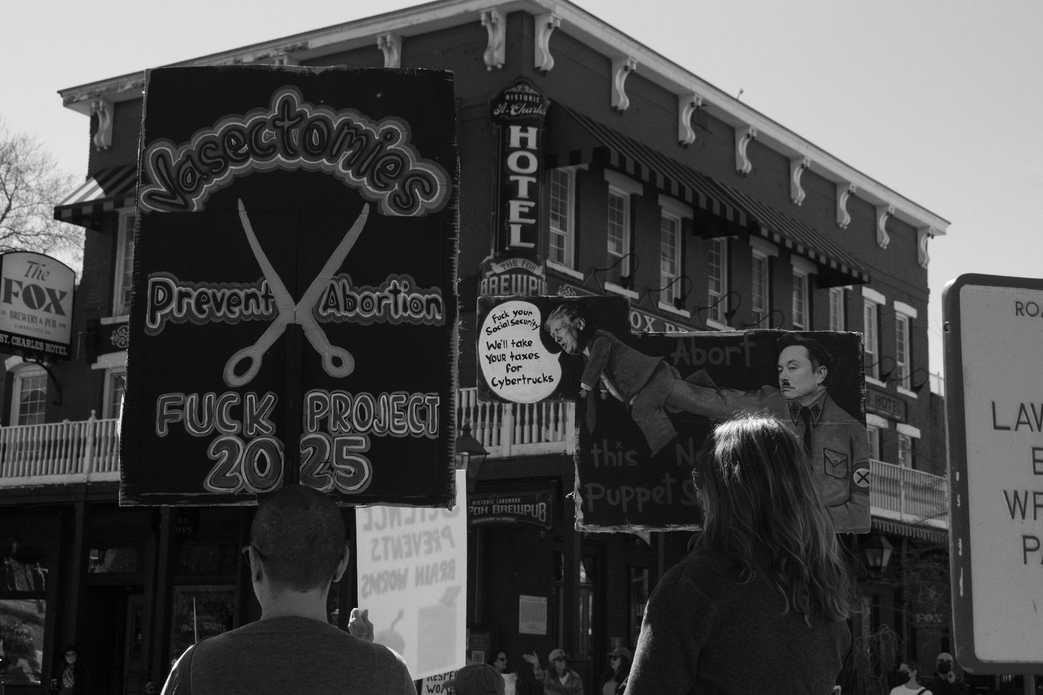 Black and white photo of protesters holding signs including anti-Project 2025 and pro-abortion rights placards at a street rally.
