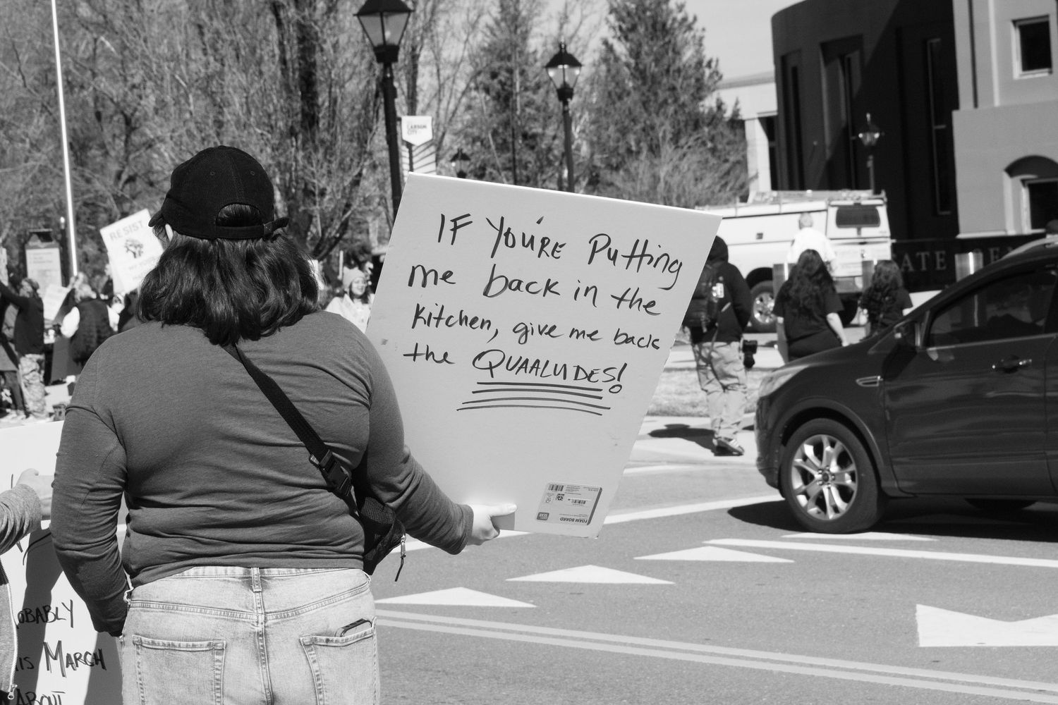 Woman holds protest sign reading If Youre Putting Me Back in the Kitchen Give Me Back the Finances at outdoor rally.