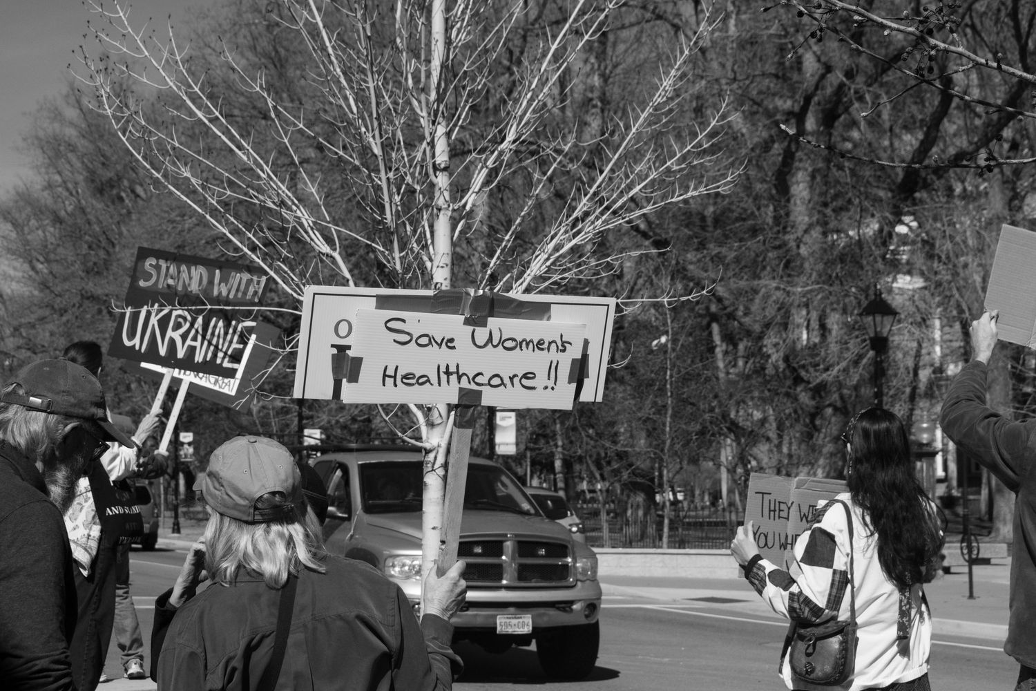 Protesters hold signs including Save Womens Healthcare and Stand With Ukraine at an outdoor street demonstration.