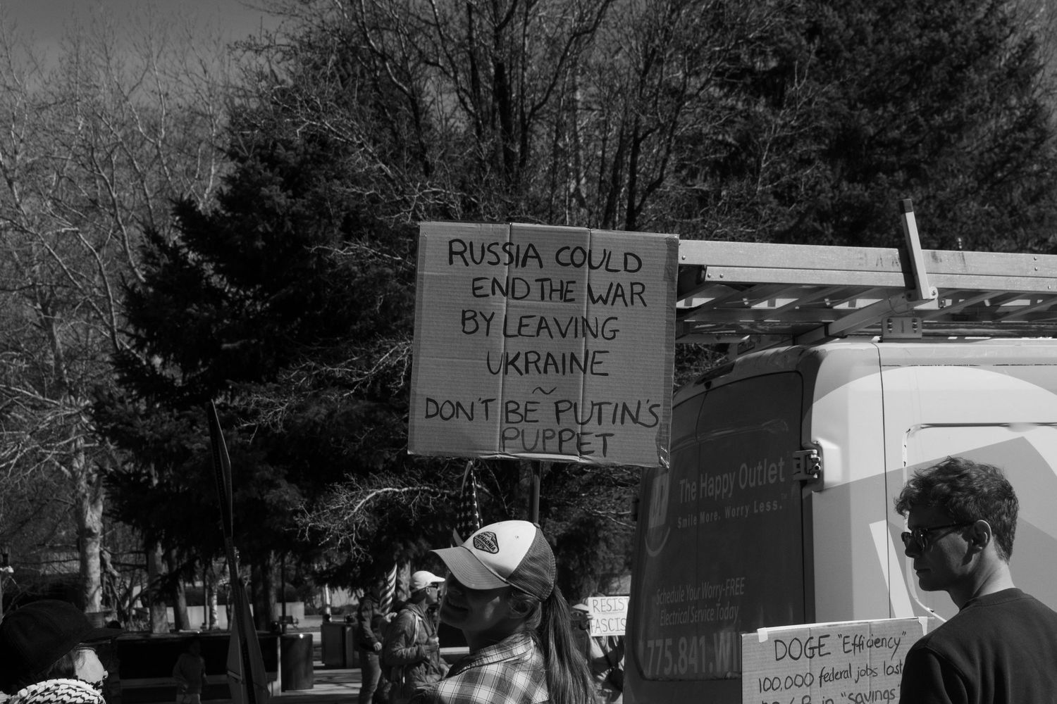 Protester holds sign reading Russia Could End the War by Leaving Ukraine, Dont Be Putins Puppet at outdoor rally.