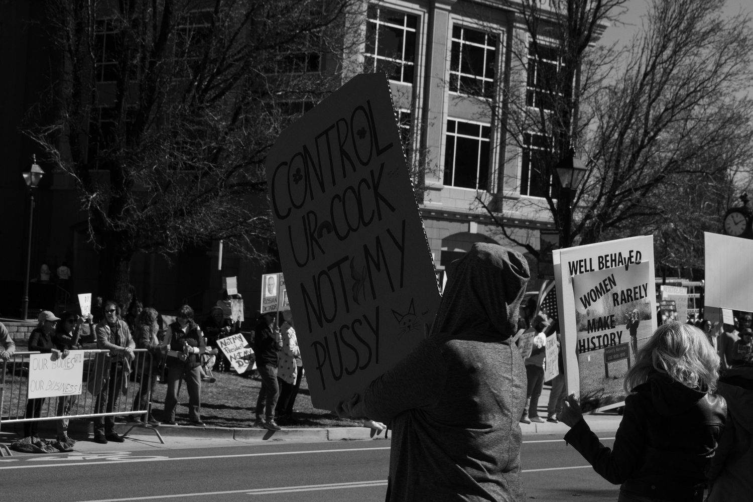 Protester holds sign reading Control Your Own Body at My Pussy during an outdoor rally near a government building.