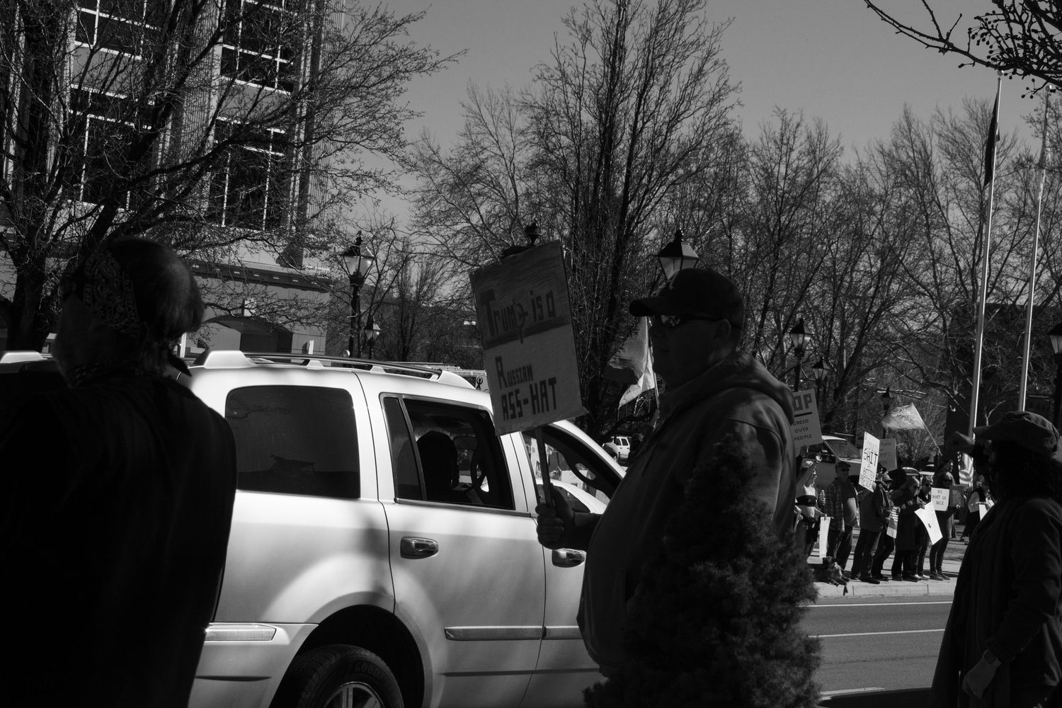 Black and white photo of protesters lining a street with signs as a white SUV passes during a daytime demonstration.