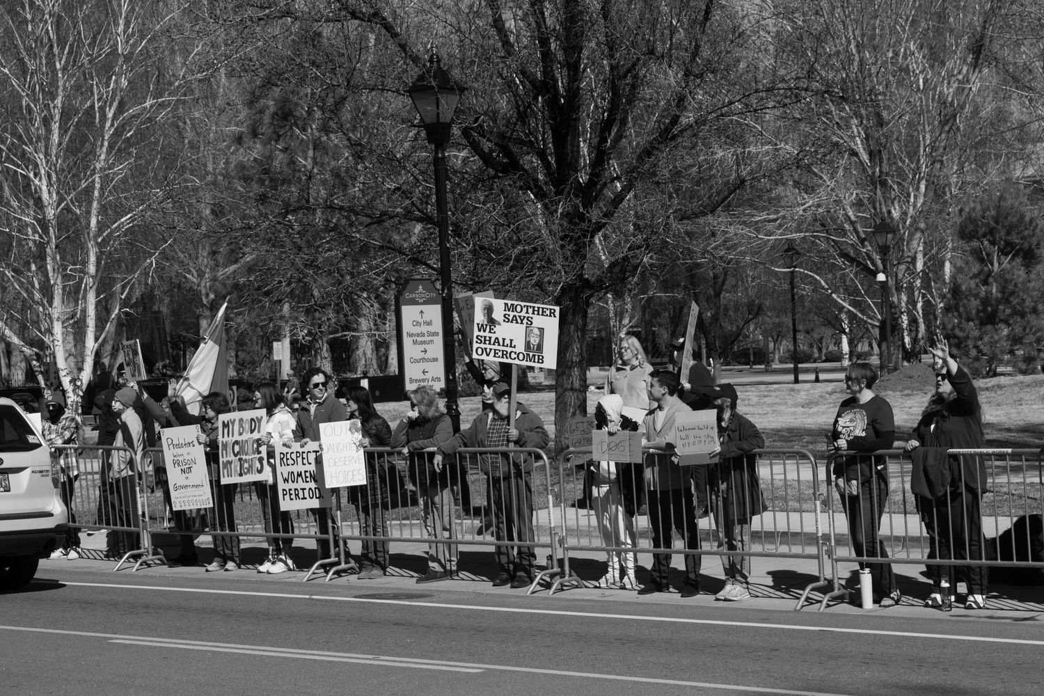 Row of protesters holding various signs stand along metal barricades on a tree-lined street during a daytime rally.