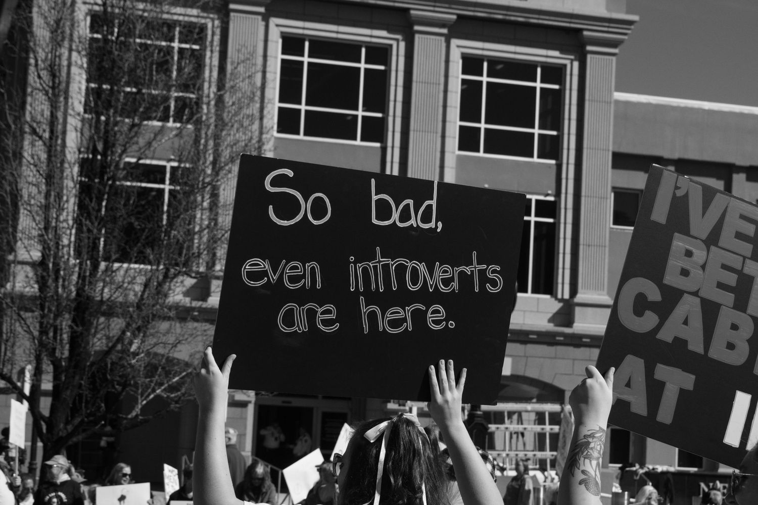 Black and white photo of protest sign reading So bad even introverts are here at a public rally.