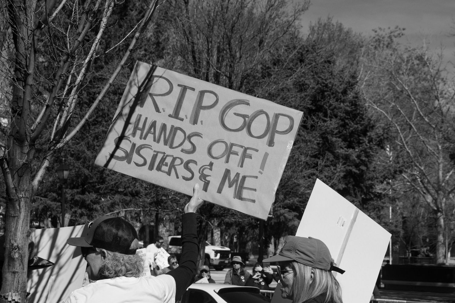 Black and white protest photo with sign reading RIP GOP Hands Off Sisters and Me at outdoor rally.
