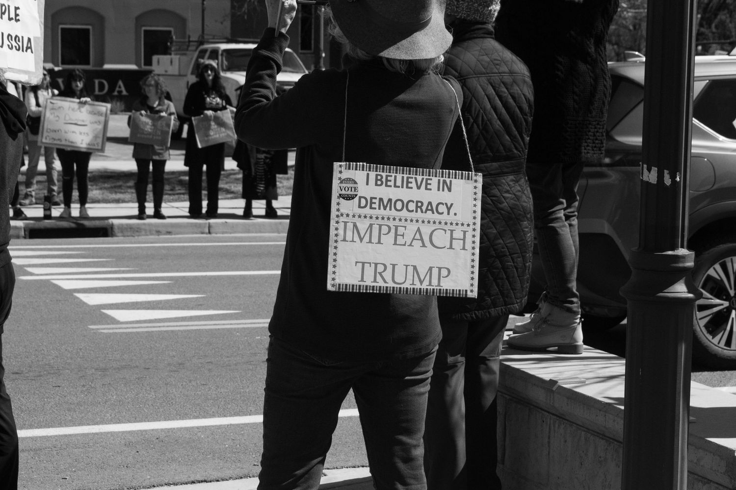 Protester wearing sandwich board sign reading I Believe in Democracy Impeach Trump at outdoor rally.
