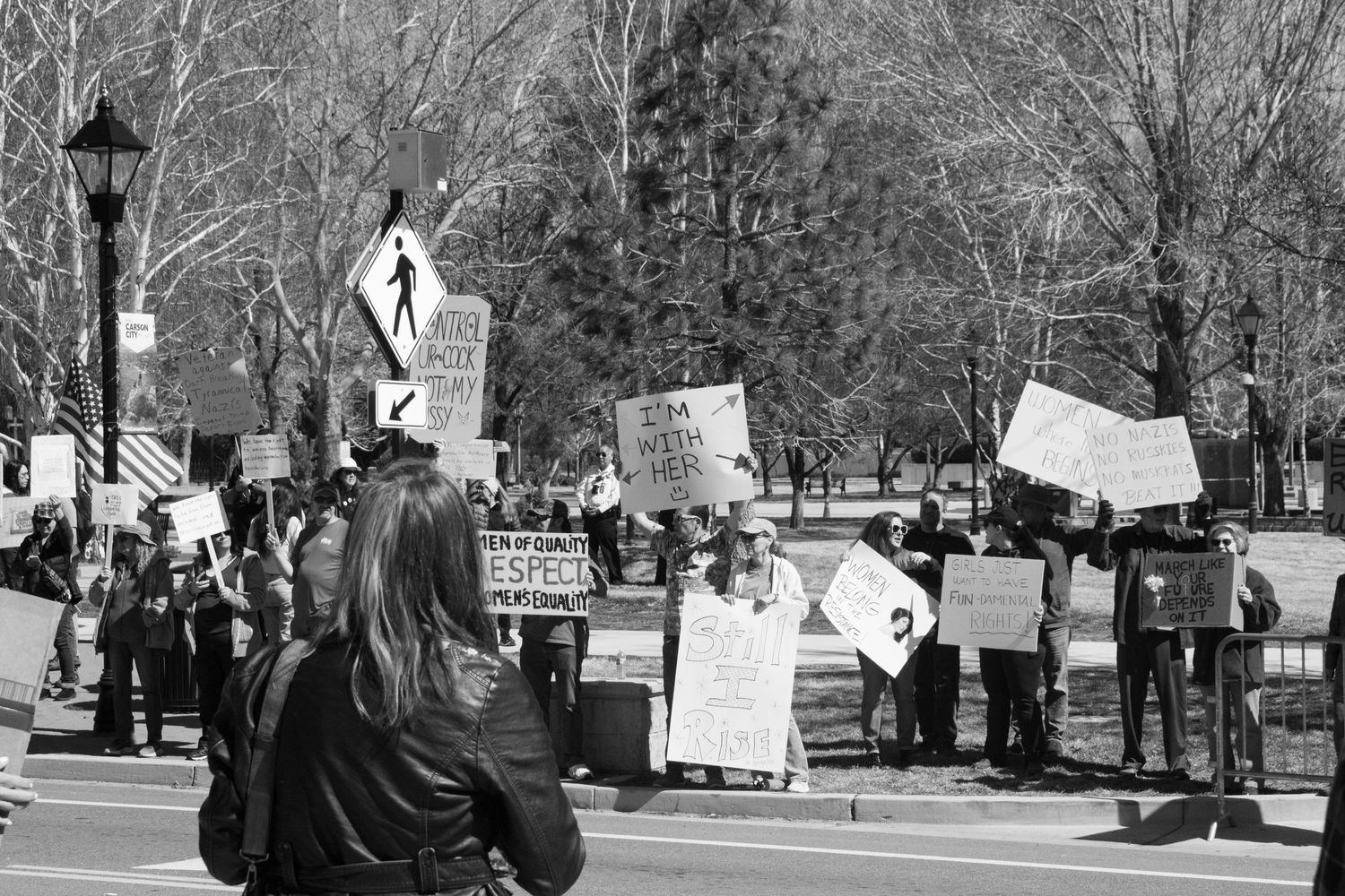 Woman photographs large group of protesters holding signs along a tree-lined street at a demonstration.