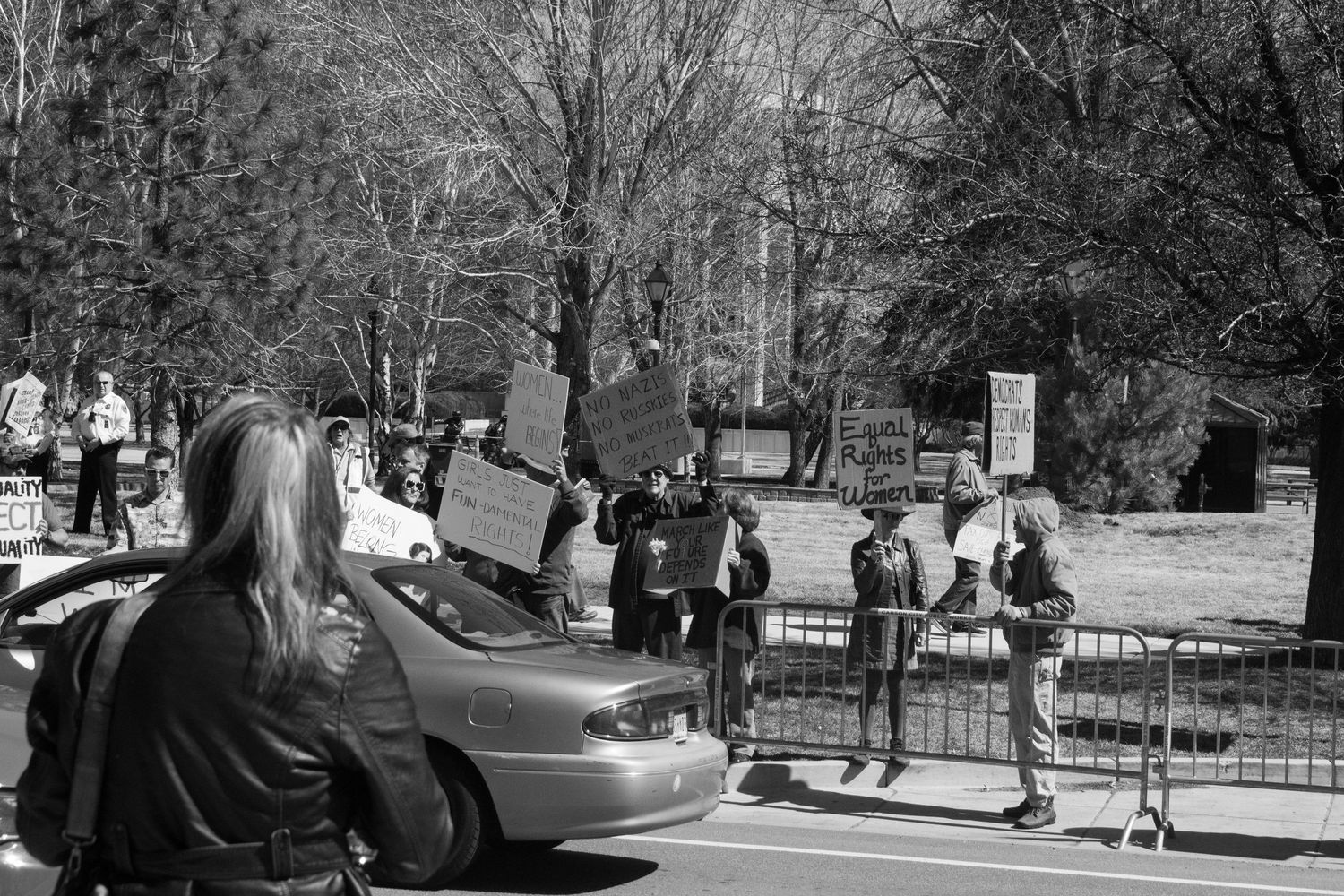 Protesters holding signs stand behind metal barricades as a car passes during an outdoor demonstration.