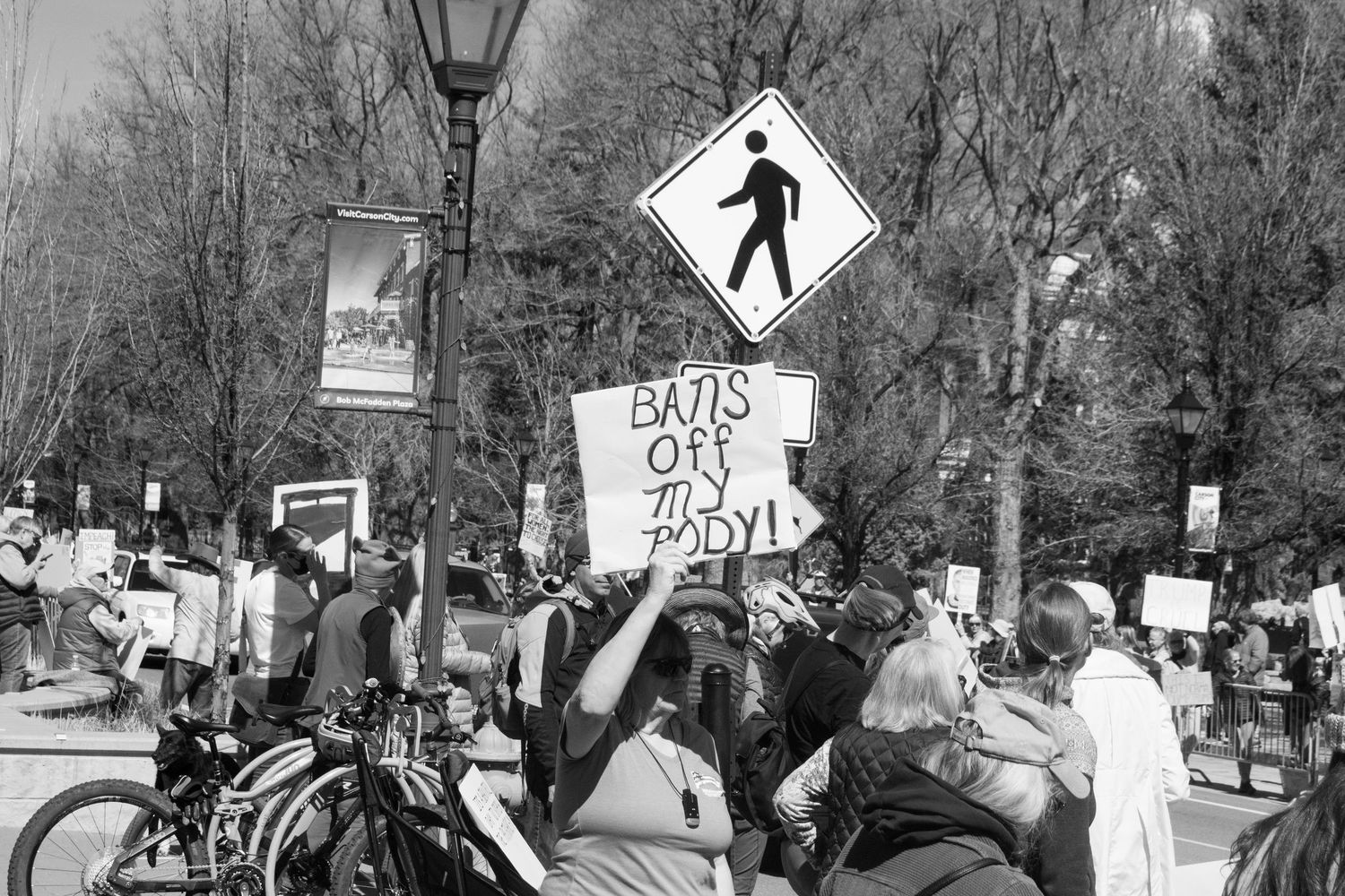Protesters hold Bans Off My Body sign beneath pedestrian crossing street sign at outdoor demonstration.