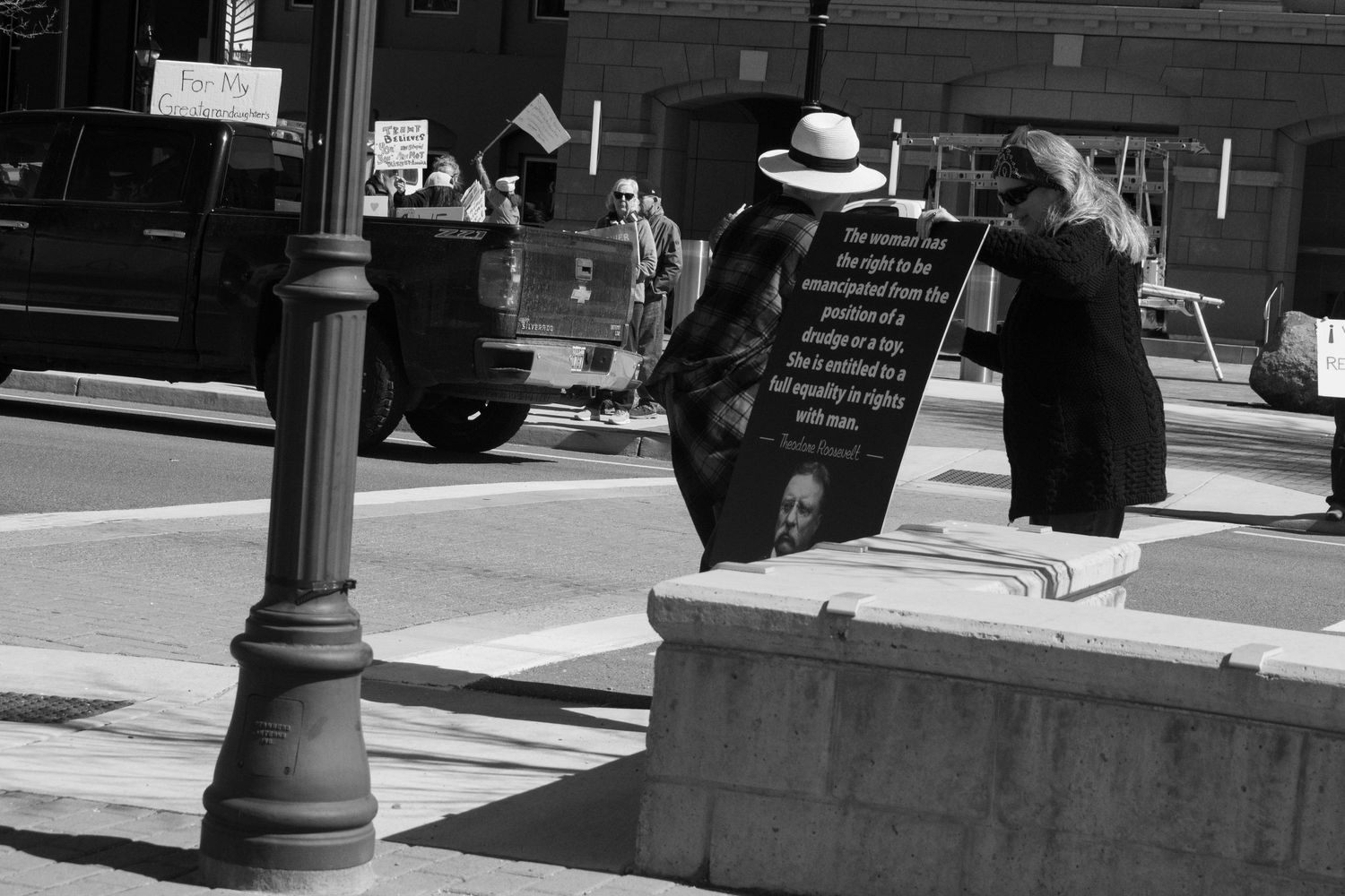 Black and white photo of two people examining a large protest sign on a city sidewalk.