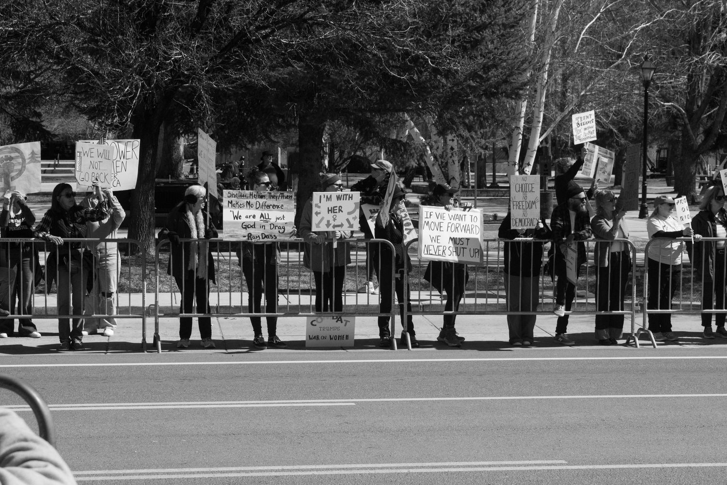 Row of protesters holding signs behind metal barriers along street in black and white photo.