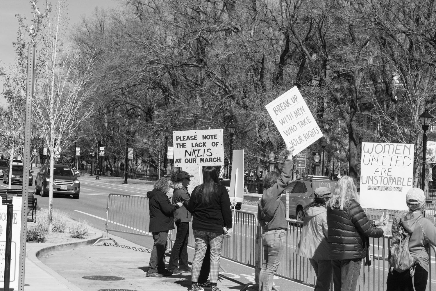 Small group of demonstrators holding protest signs along barricaded sidewalk in black and white.