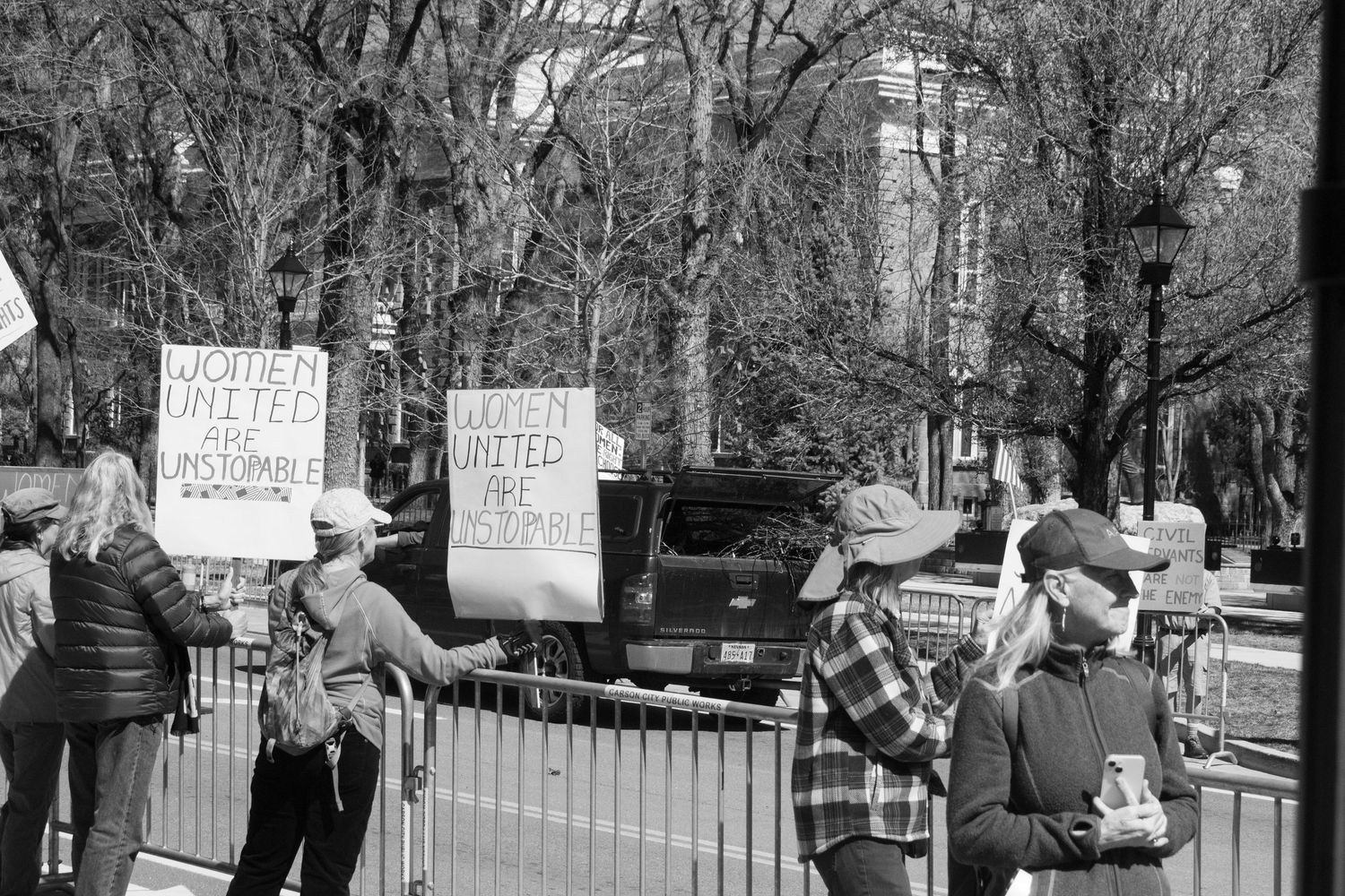 Protesters hold Women United Are Unstoppable signs along metal barrier fence at outdoor rally.