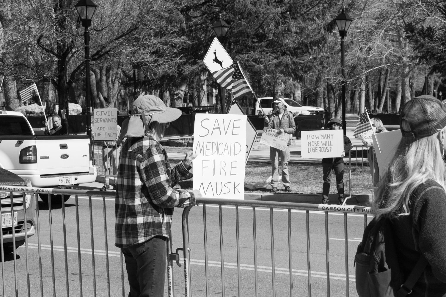 Protester holds Save Democracy Fire Musk sign near metal barricade at outdoor demonstration.