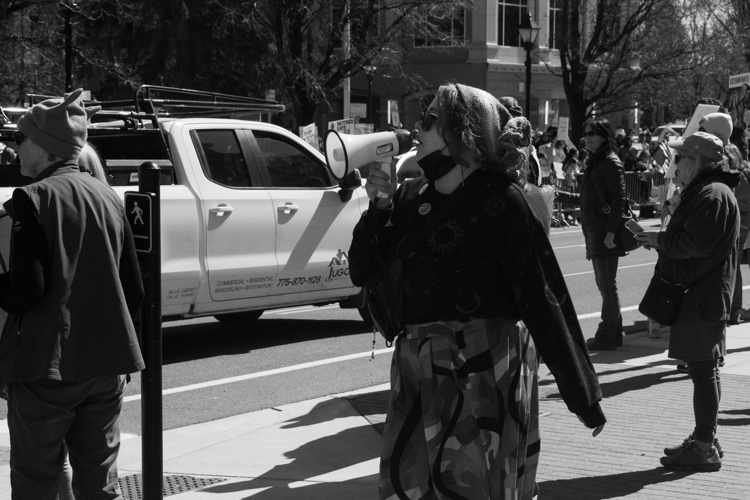 Woman speaks into megaphone wearing camouflage pants at outdoor street protest demonstration.