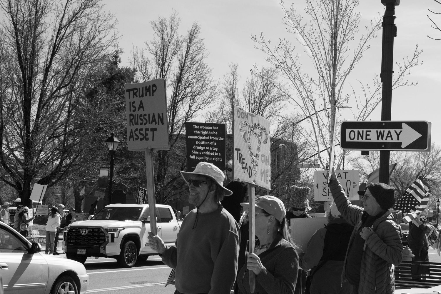 Black and white protest photo showing demonstrators holding a sign reading Trump Is A Russian Asset near a One Way street sign.