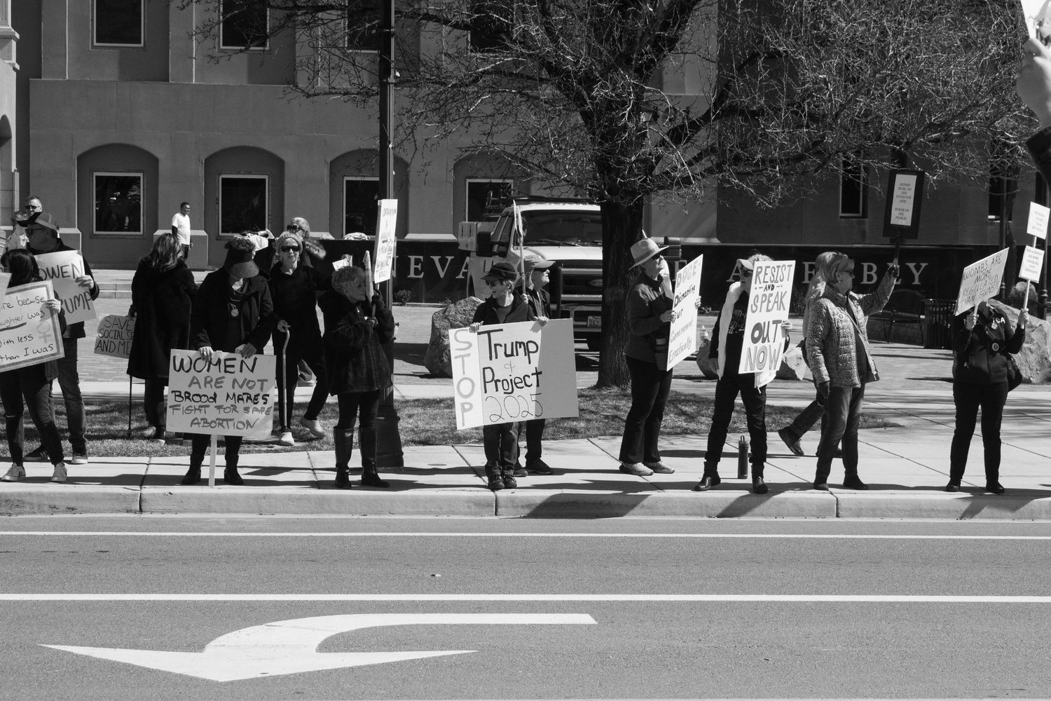 Black and white photo of protesters lining a sidewalk holding various signs including Women's Rights and Trump Project.