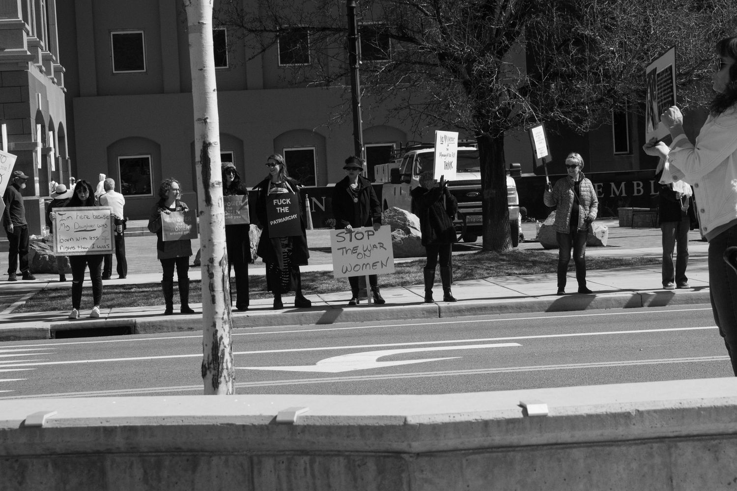 Black and white photo of protesters standing along a sidewalk holding signs at a public demonstration.