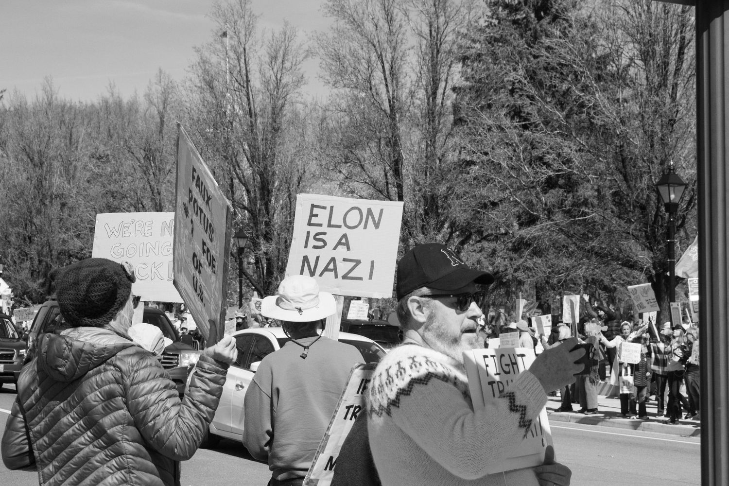 Black and white image of protesters at a rally holding a sign reading Elon Is A Nazi amid a crowd of demonstrators.