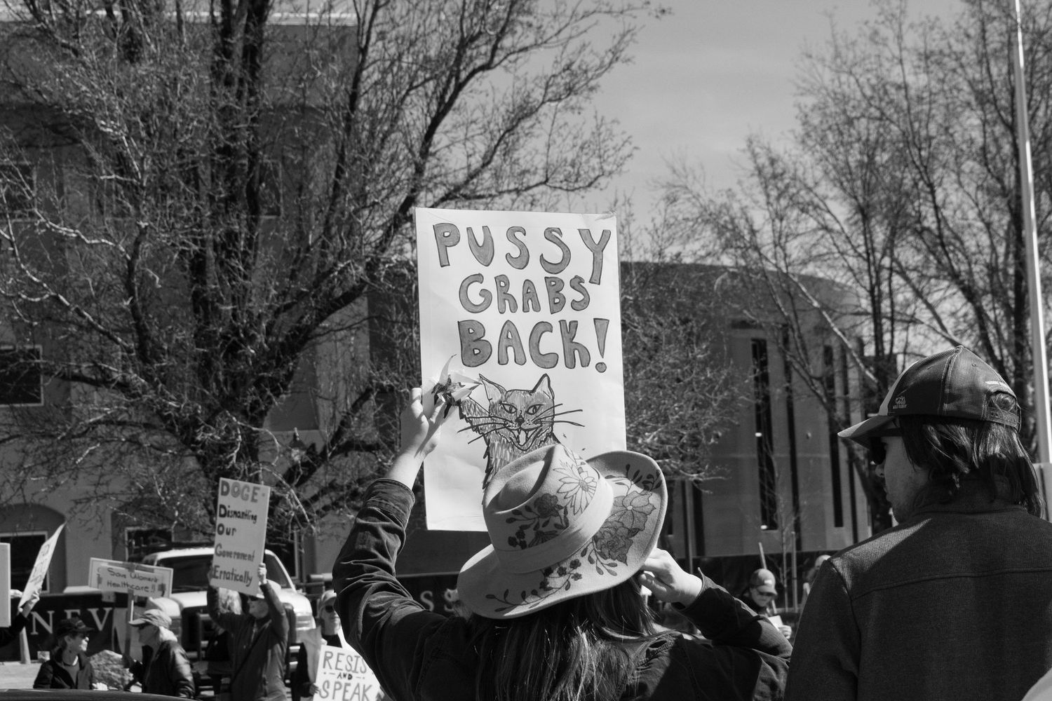 Black and white protest photo featuring a demonstrator holding a sign reading Pussy Grabs Back with a cat illustration.