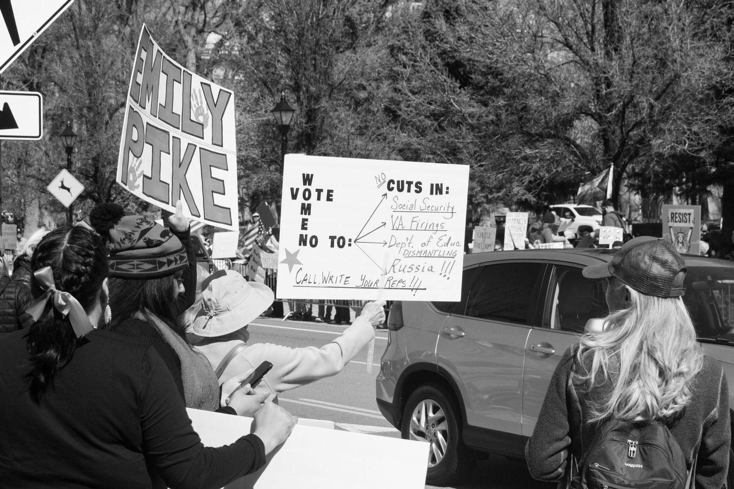 Black and white photo of protesters holding signs including Holy Mike and Cuts In Vote No at an outdoor demonstration.