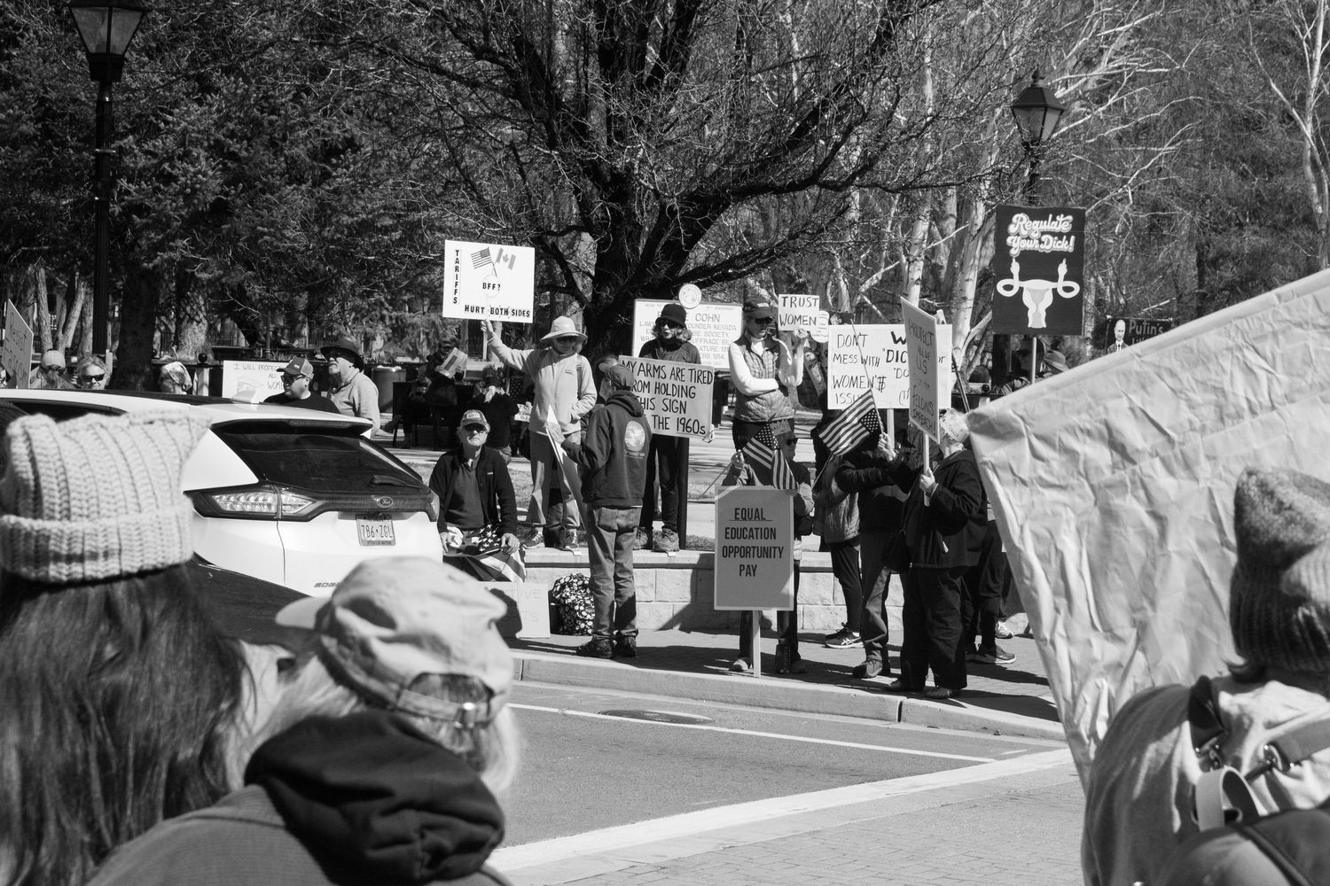 Black and white wide shot of protesters carrying signs along a stone wall barrier at an outdoor political rally.