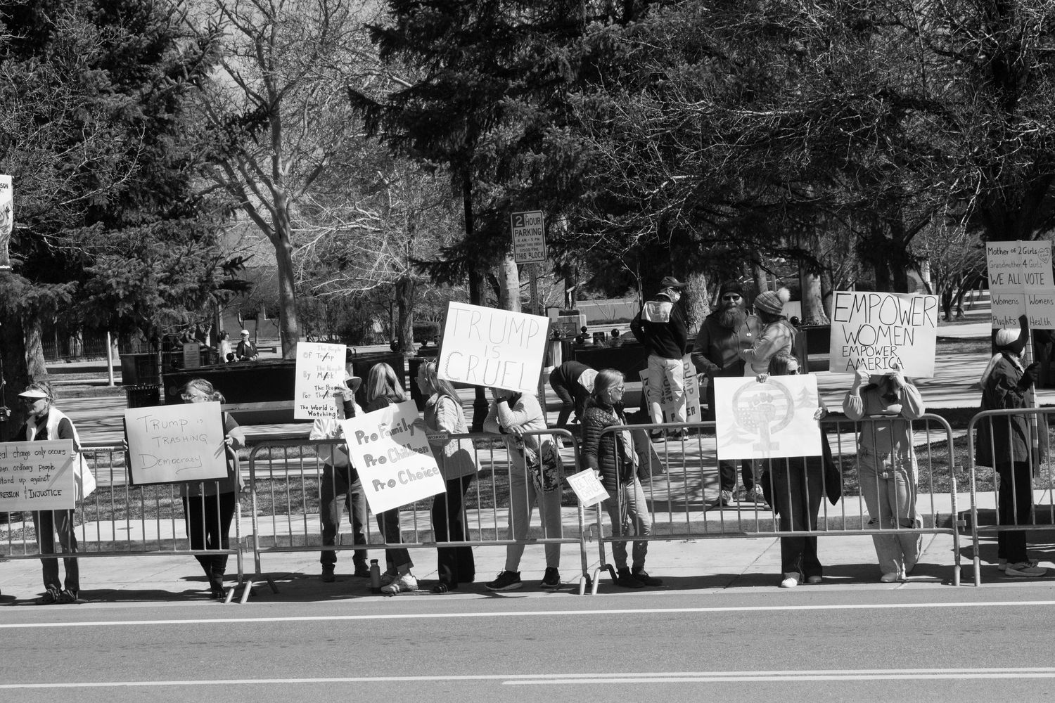 Black and white photo of a line of protesters holding handmade signs along a street barrier at an outdoor demonstration.
