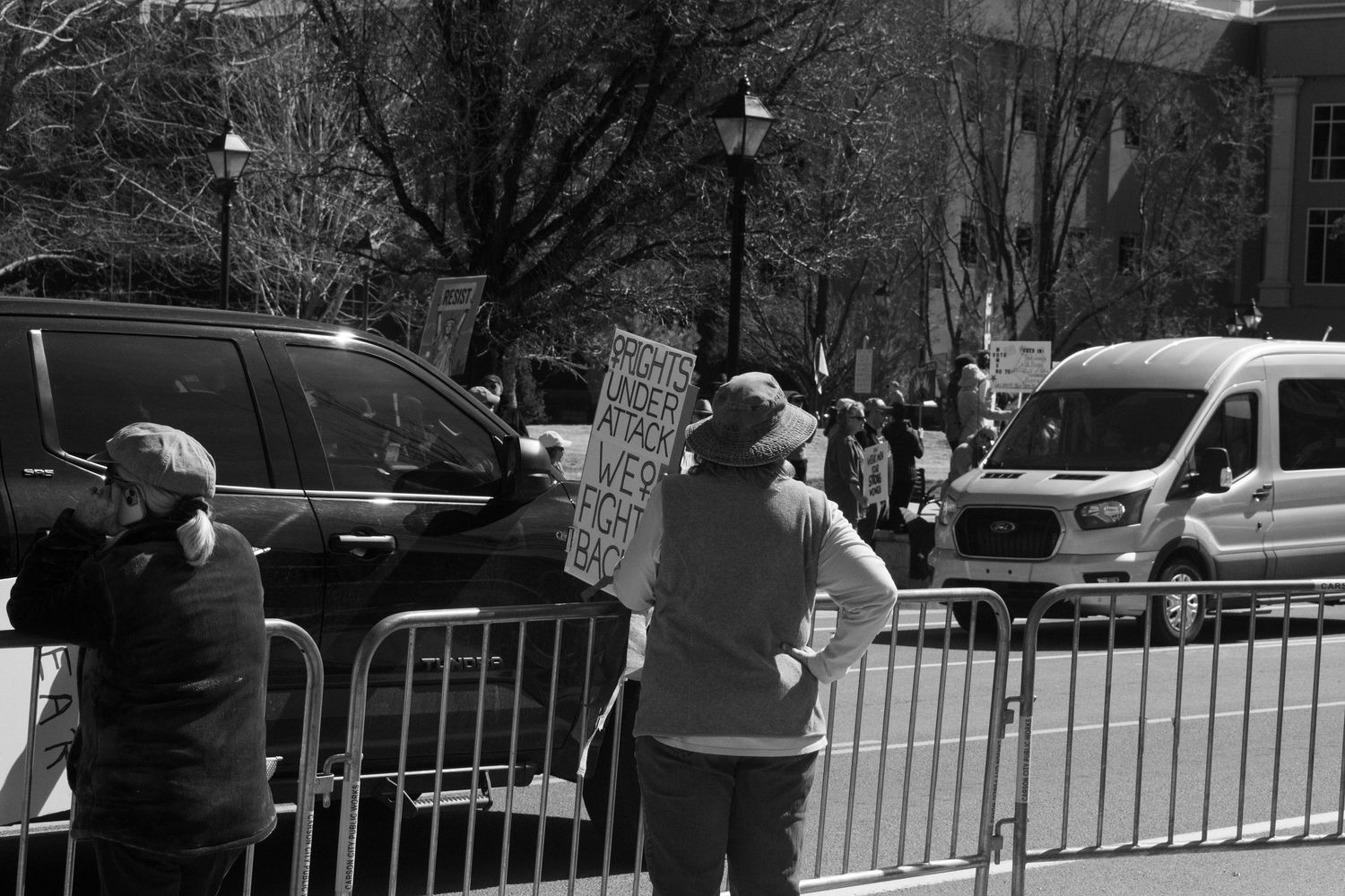 Black and white photo of protesters holding signs near metal barriers on a city street.