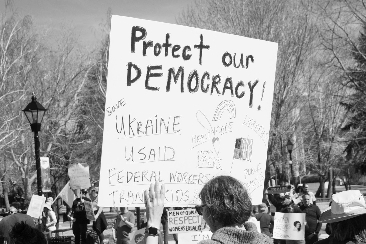 Protest sign reading Protect Our Democracy Save Ukraine USAID Federal Workers Trans Kids.