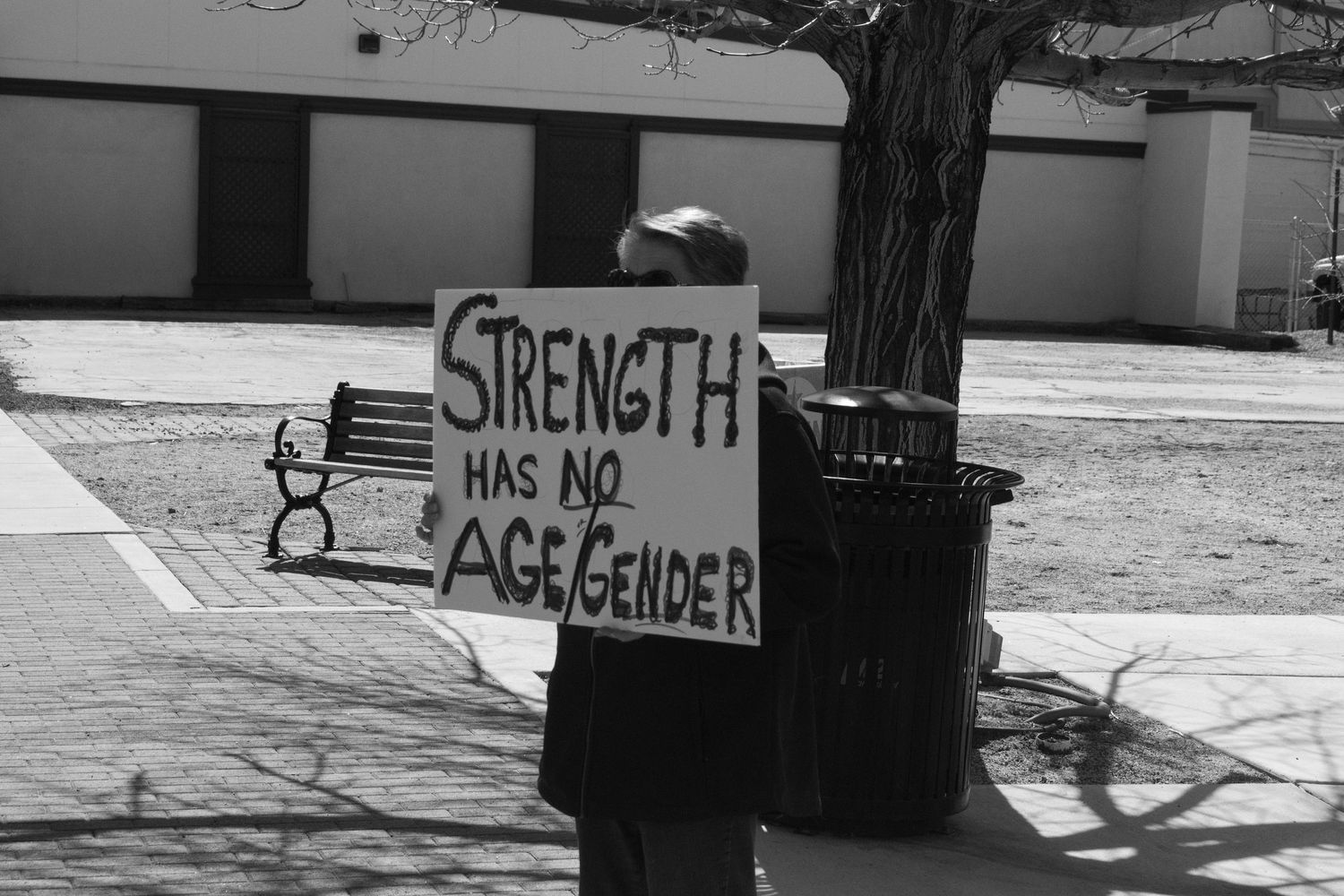 Person holding a cardboard sign reading Strength Has No Age Gender near a tree.